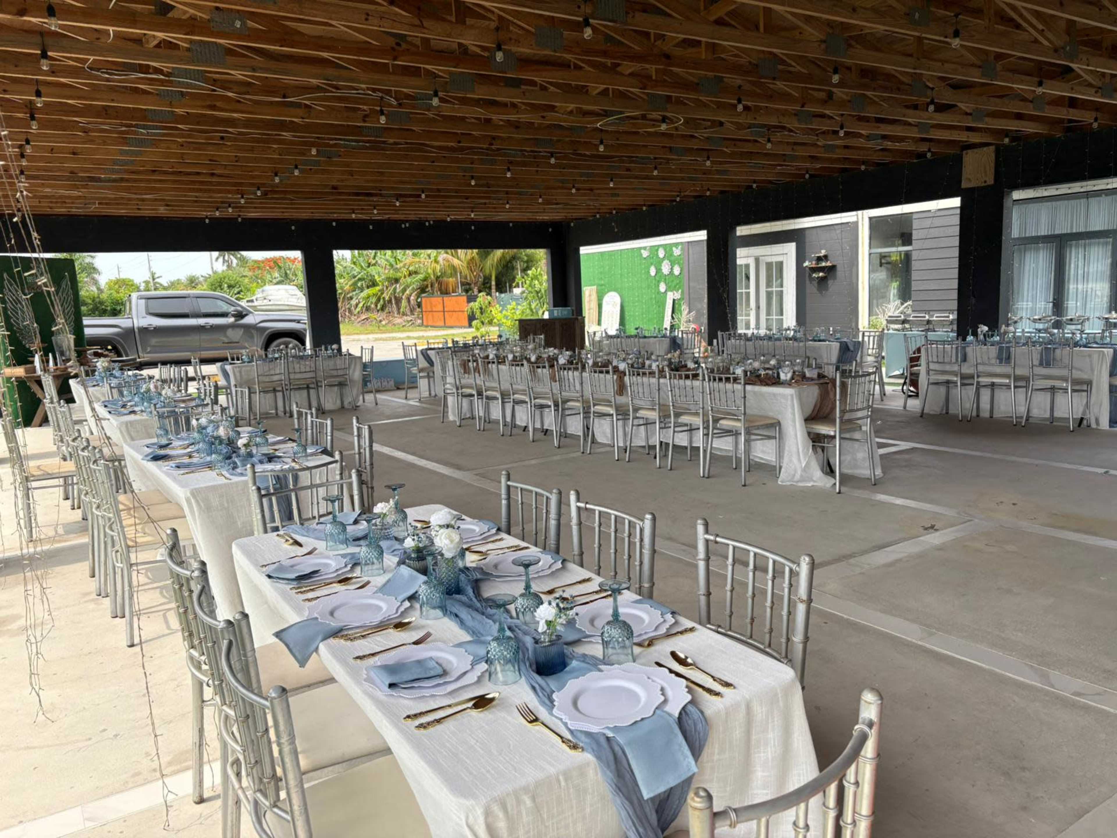 A large outdoor dining area is set up with elegantly arranged tables covered in white linens, each featuring place settings of glassware and utensils, under a wooden roof.