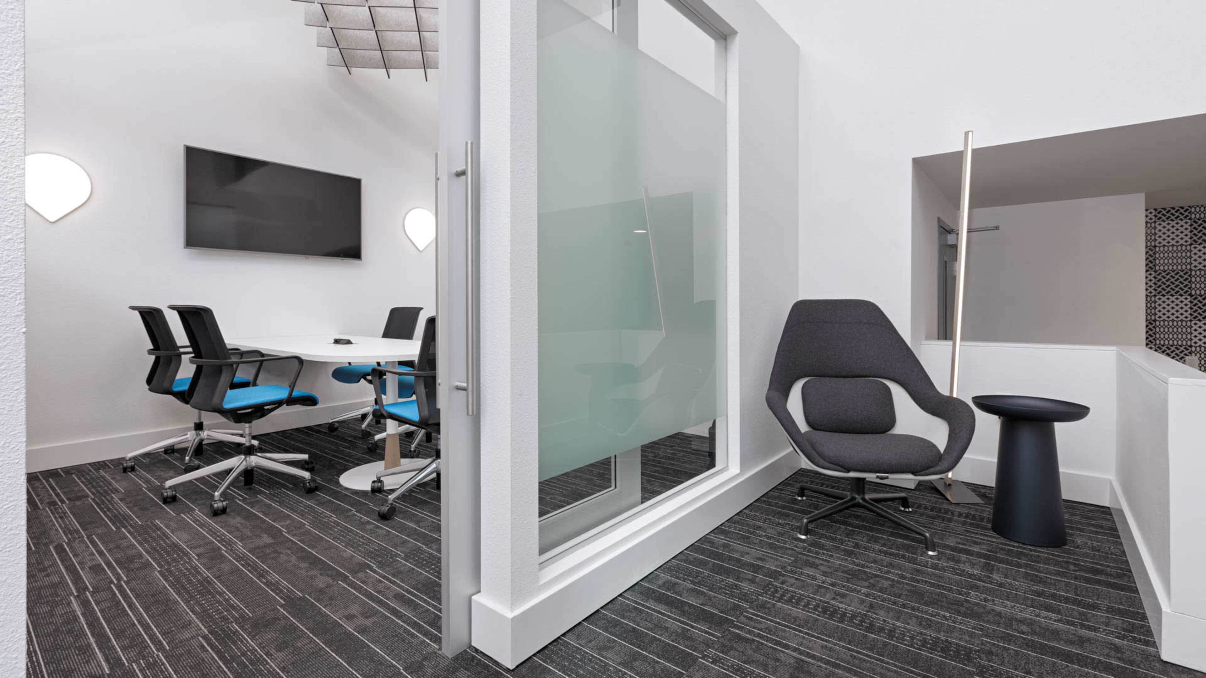 A modern conference room features a large table surrounded by ergonomic chairs, with a TV mounted on the wall and a glass partition separating it from an adjacent seating area.