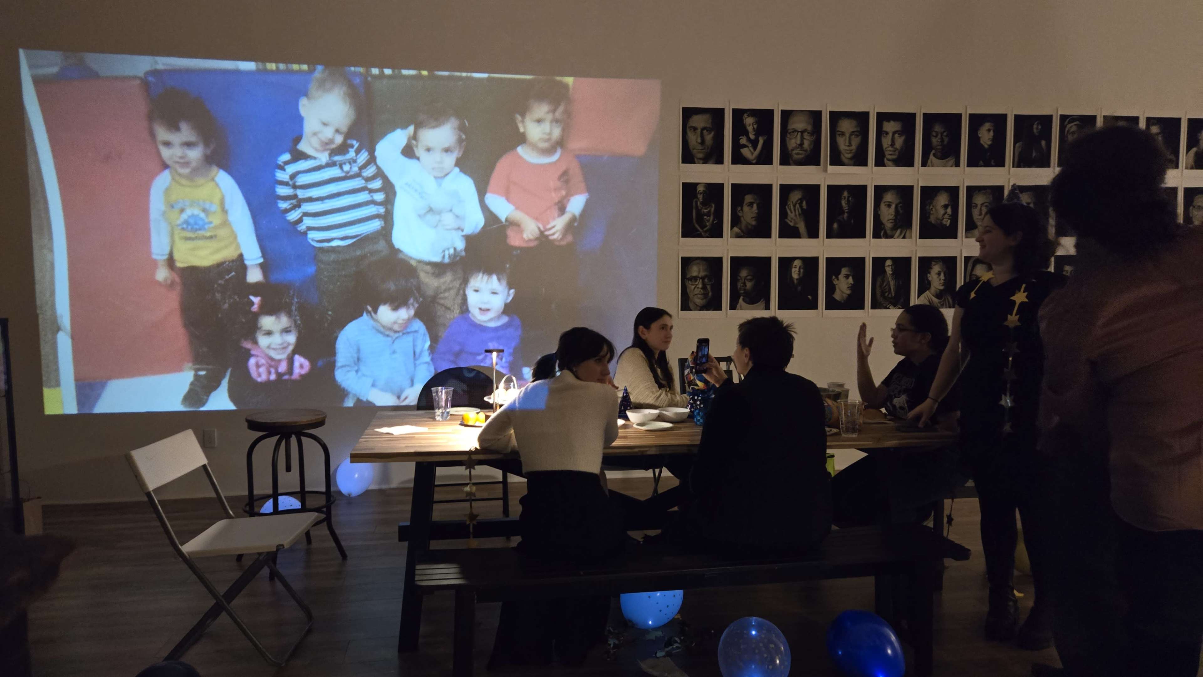 A group of people sits around a table in a room with a projector displaying a childhood photograph of several children on a wall.