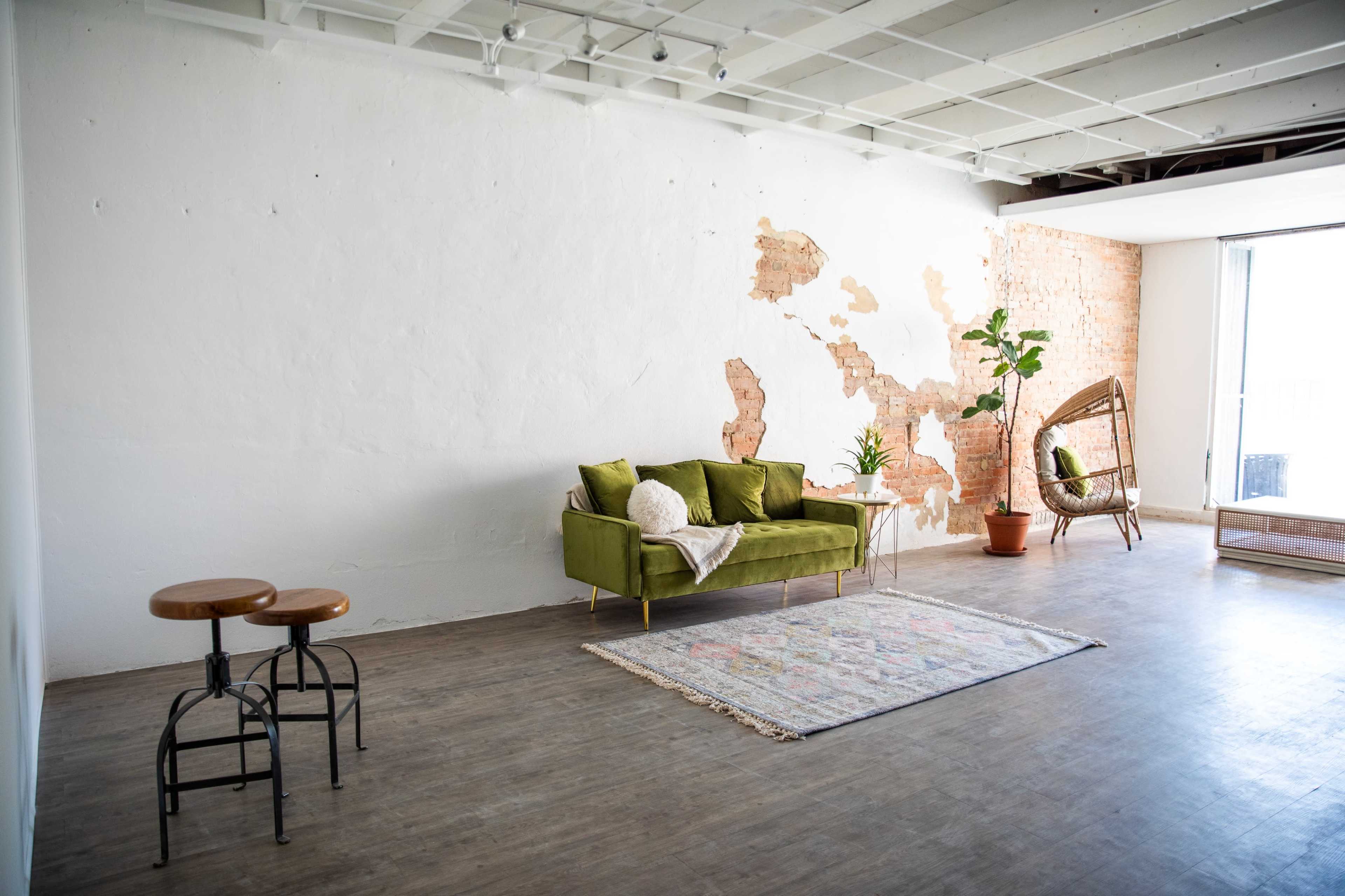 The image shows a bright, minimalist room featuring a green sofa, two stools, a patterned rug, and a wall with a subtle map design made of brick.