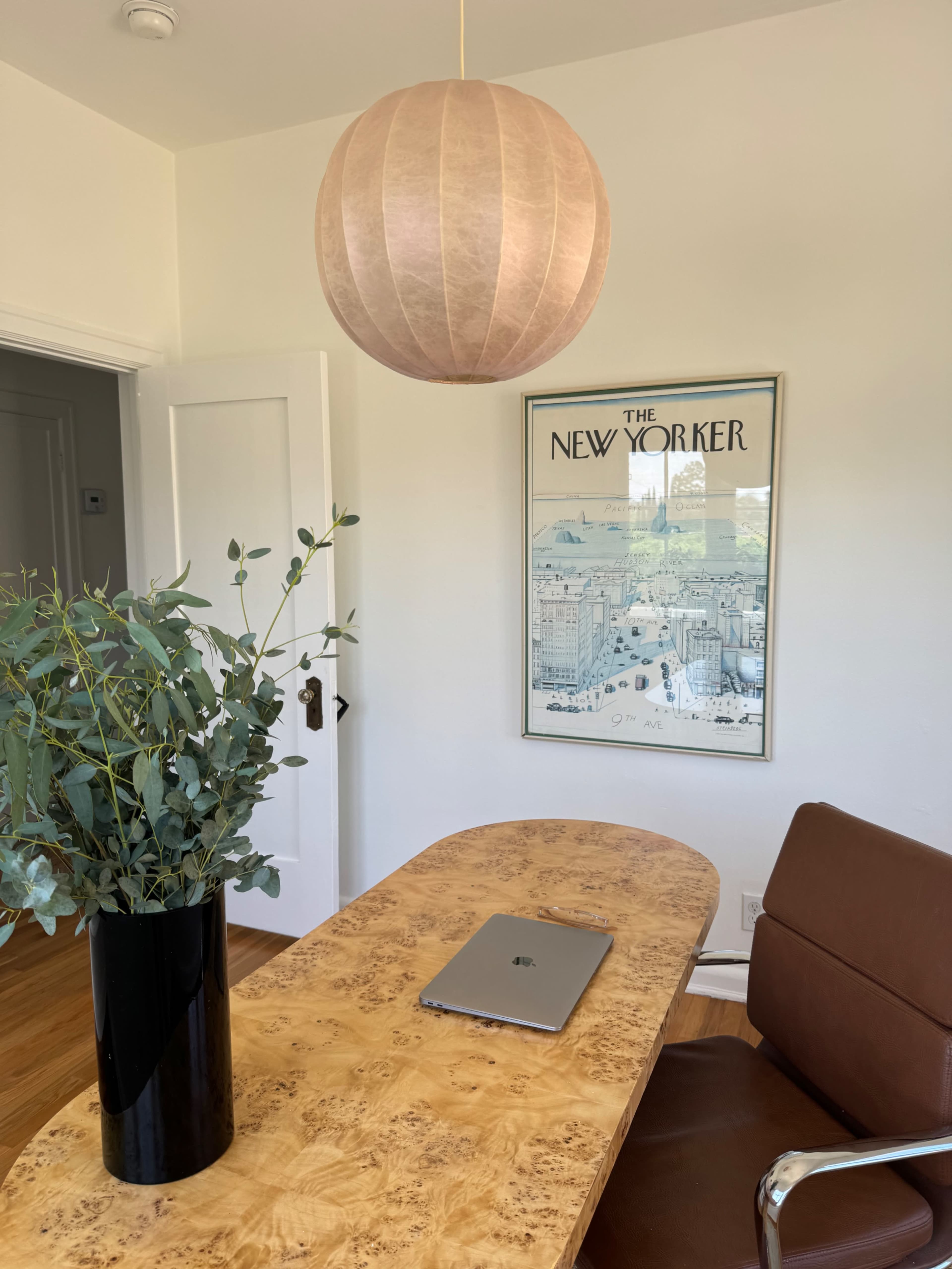 A modern workspace features a wooden table with a laptop, a tall black vase holding eucalyptus leaves, and a framed New Yorker magazine cover on the wall.