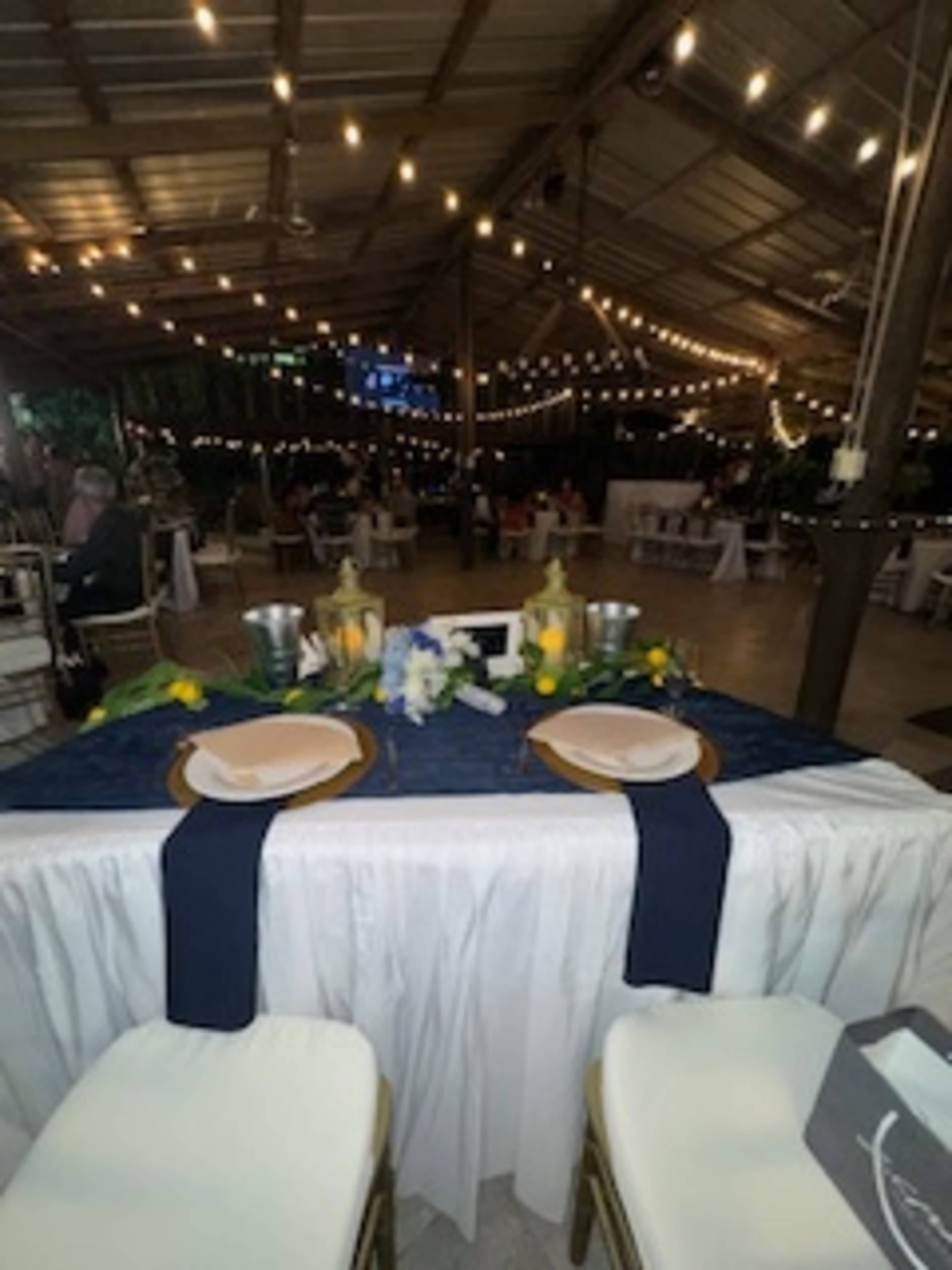 A decorated dining area features a table set for two, with floral centerpieces and string lights overhead in a large indoor space.