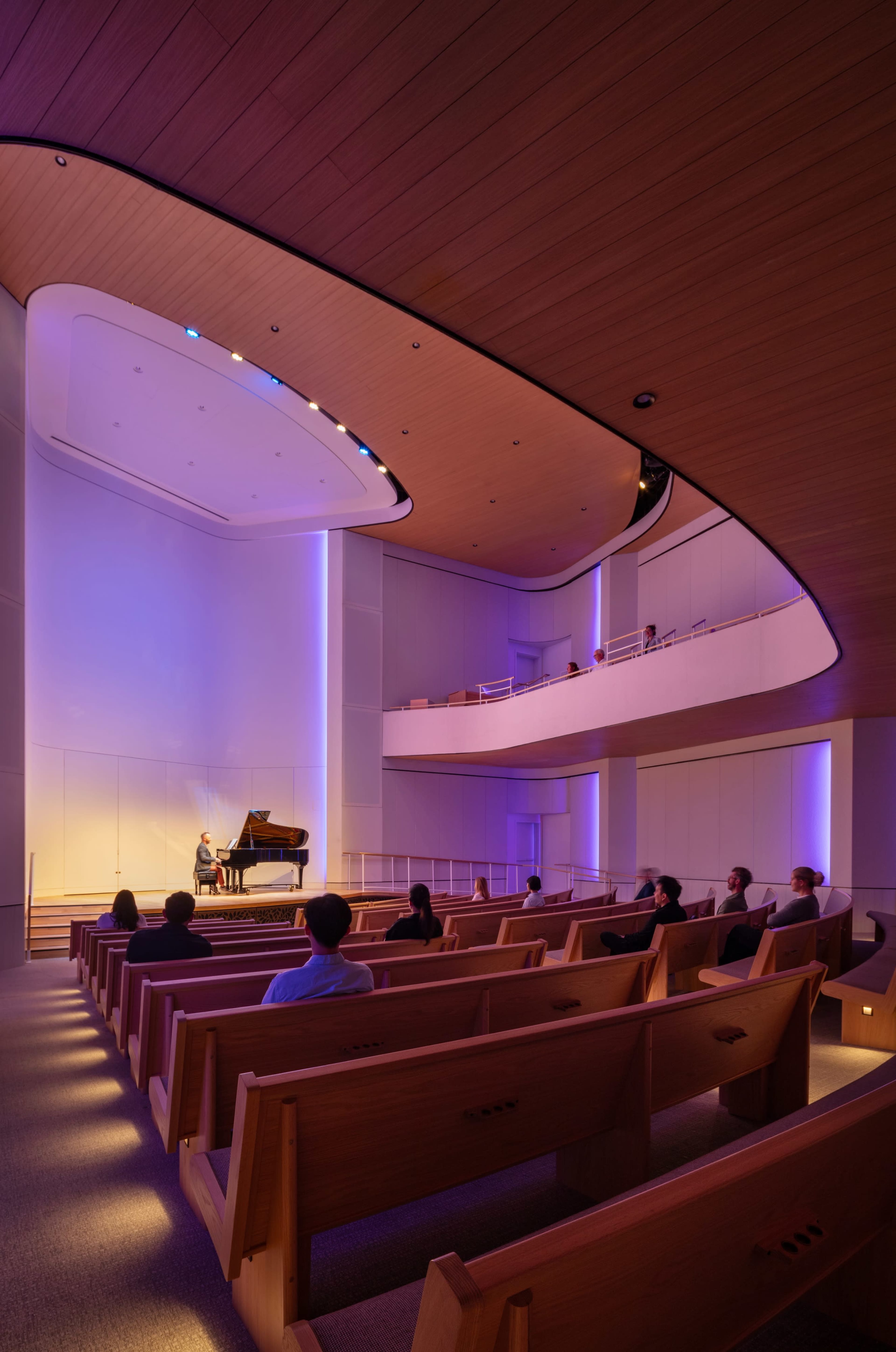 A performer plays a piano on stage in a modern concert hall with wooden seating and ambient lighting.