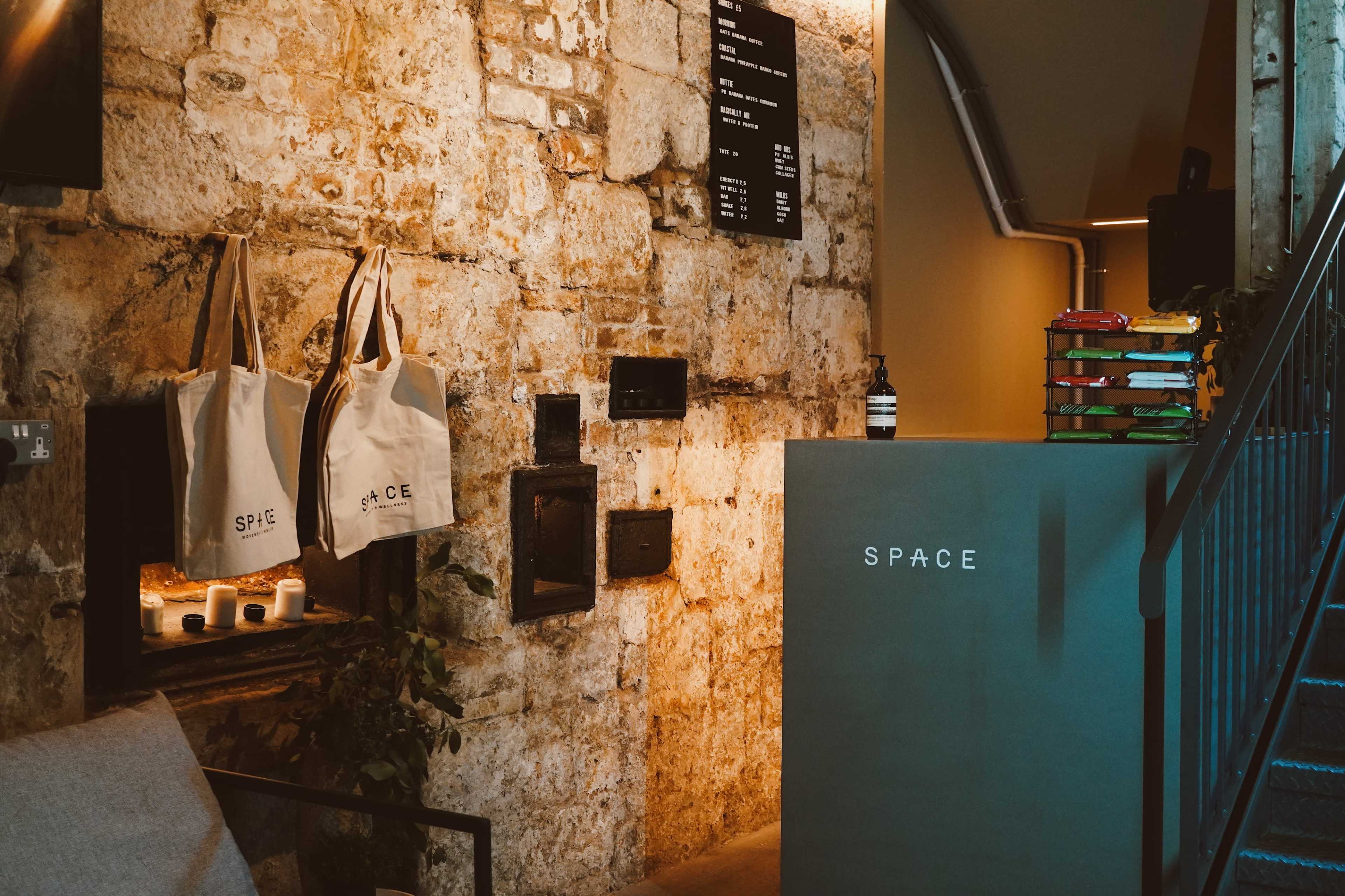 The image shows a rustic interior with a stone wall, a reception desk labeled "SPACE," and hanging tote bags displayed nearby.