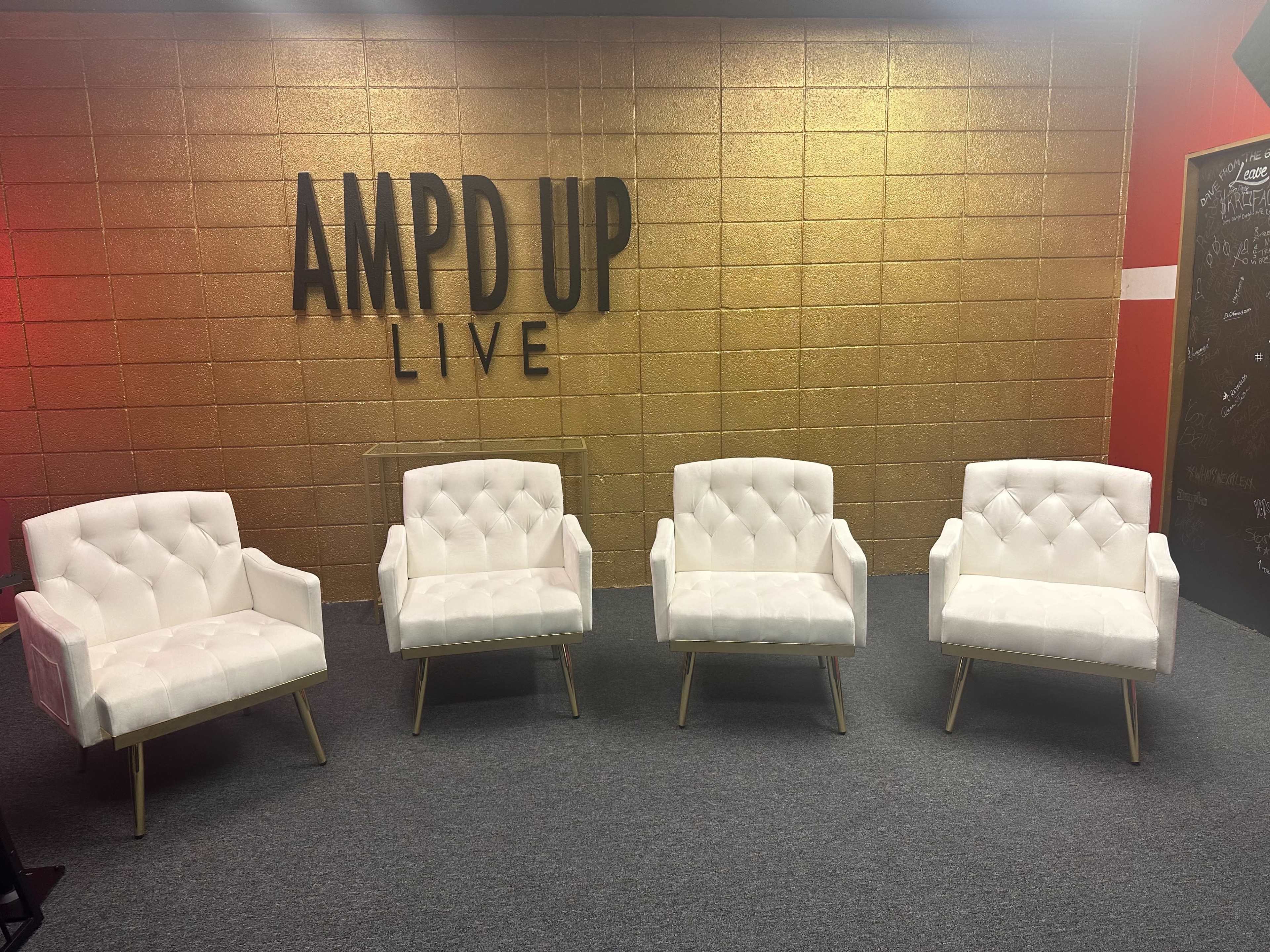 Four white tufted armchairs arranged in front of a gold and black wall with the text "AMPD UP LIVE" displayed.