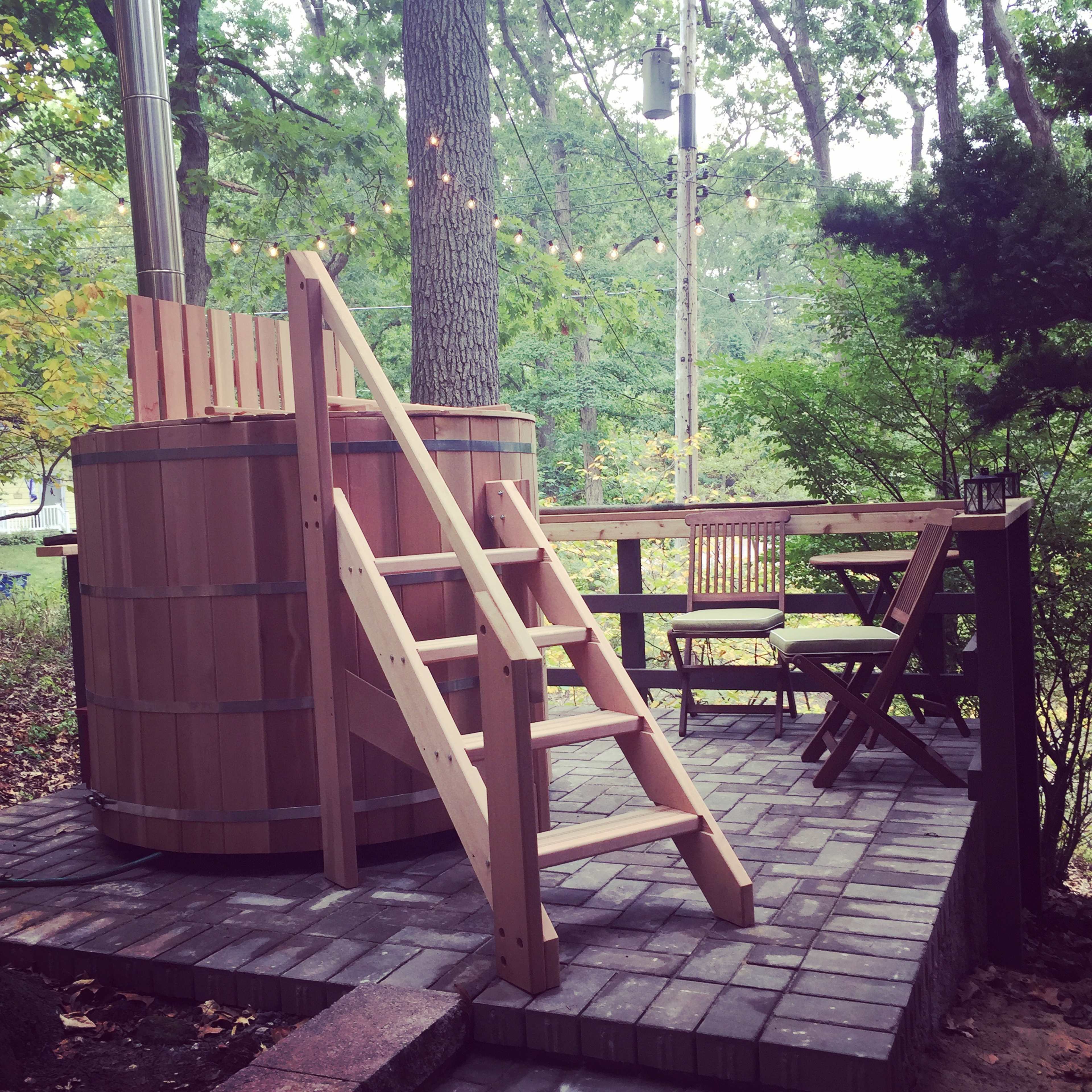 A wooden hot tub surrounded by trees, with a set of stairs leading up to it and a small seating area nearby.