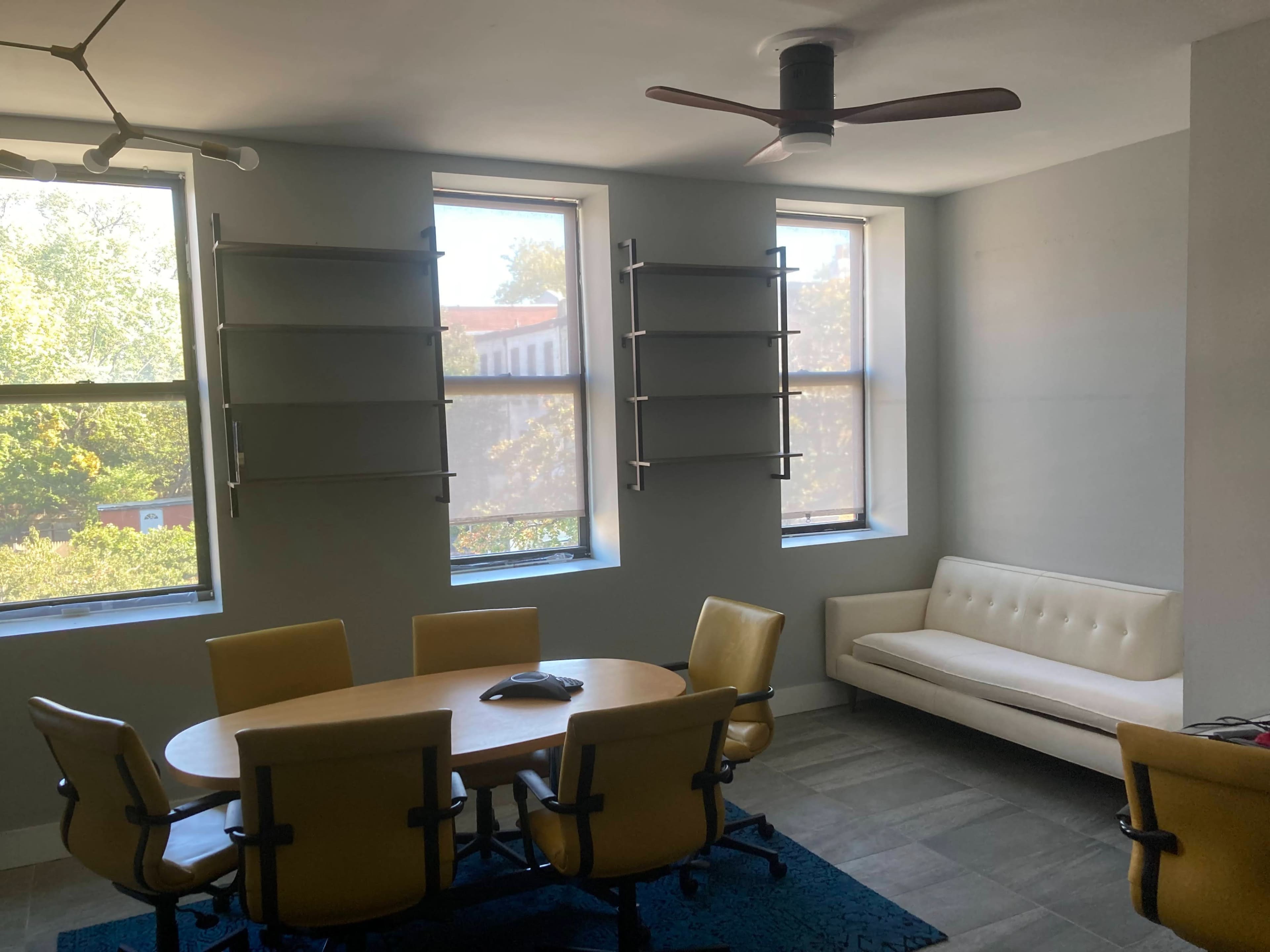 A meeting room features a round table surrounded by yellow chairs, with a white sofa and windows offering natural light.