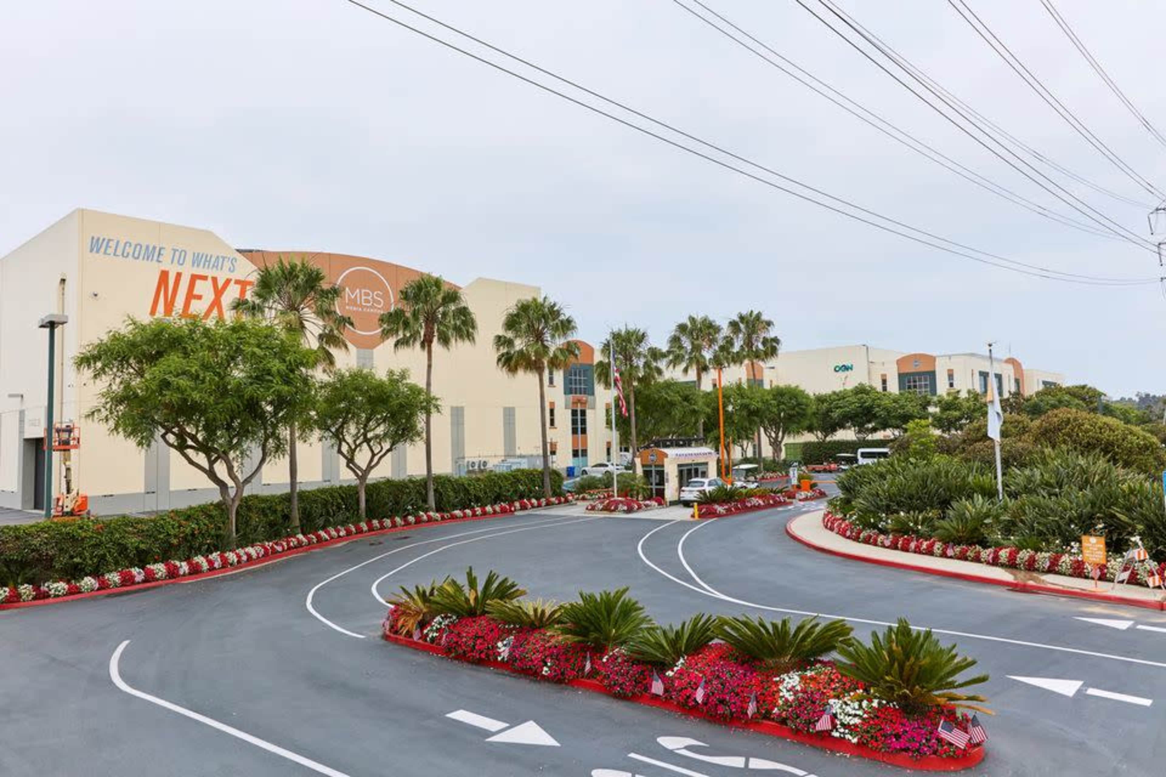 A parking lot entrance to a commercial complex with palm trees and colorful flower beds lining the curved drive.