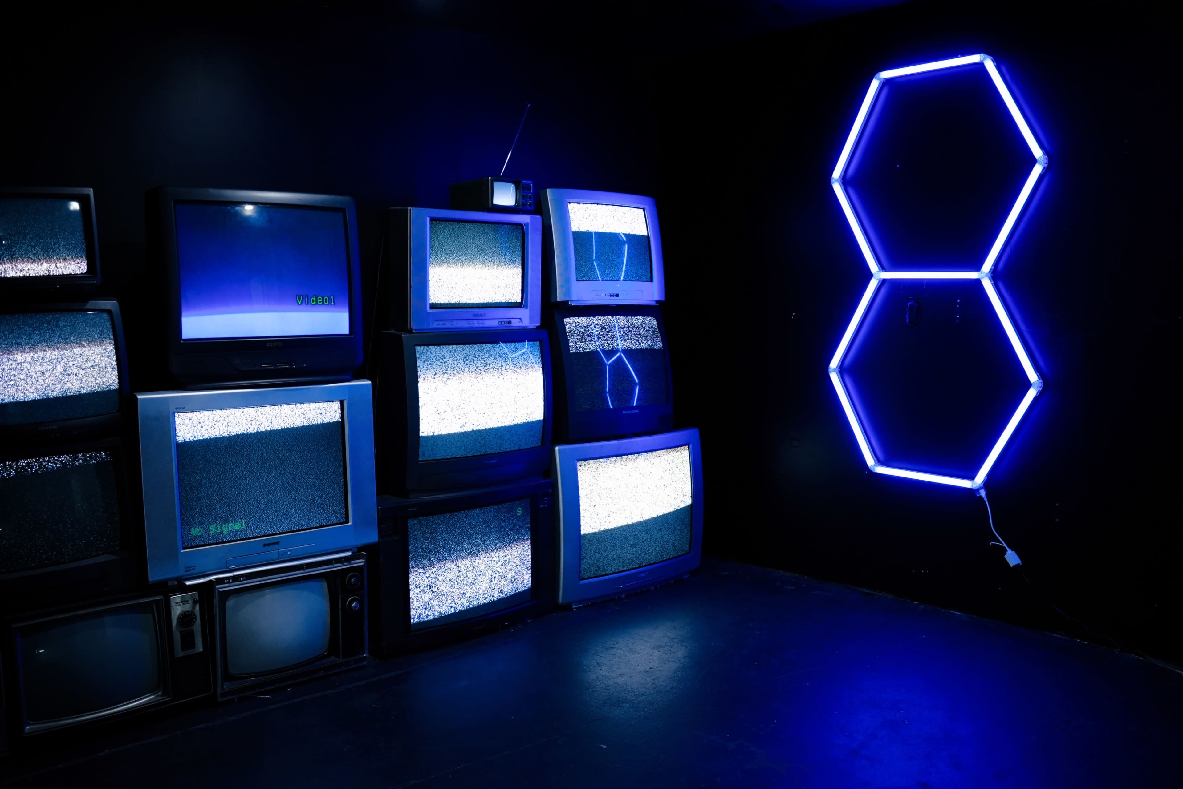 A collection of vintage televisions displaying static is arranged against a dark wall, alongside a neon blue hexagonal light fixture forming the number 8.