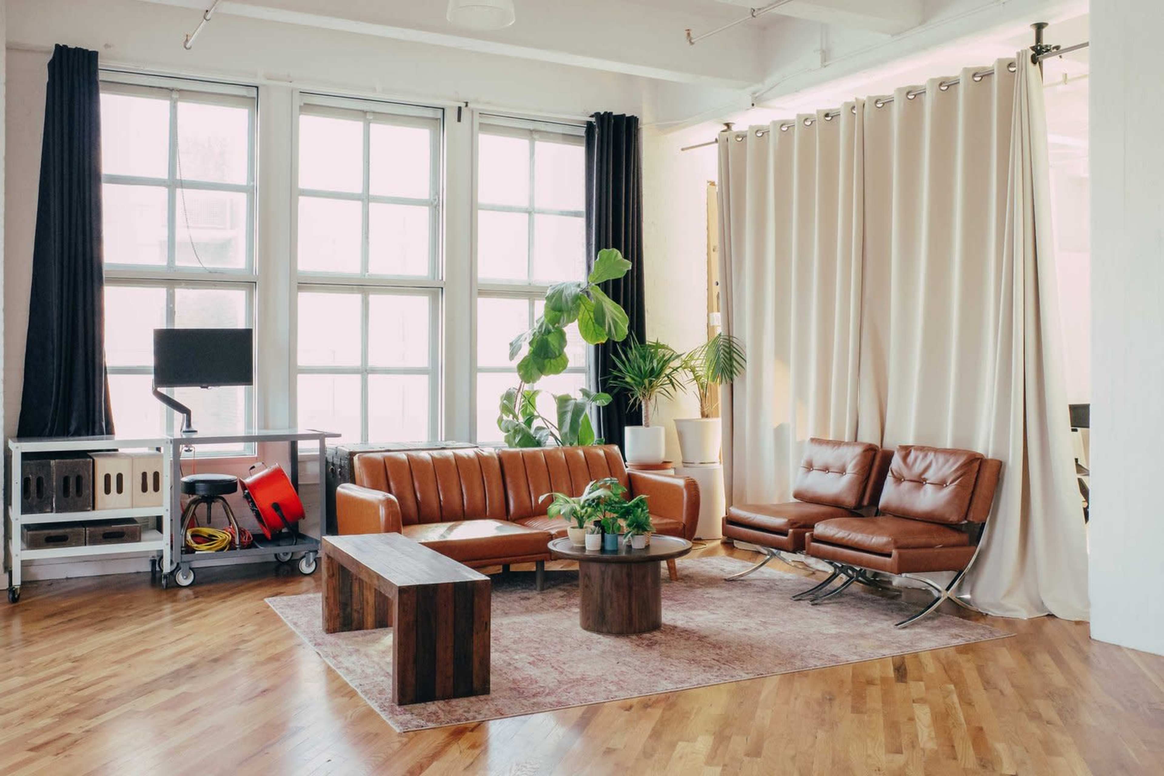 A modern living room featuring brown leather sofas, a circular wooden coffee table, and large windows with sheer curtains, complemented by potted plants and a wooden bench.