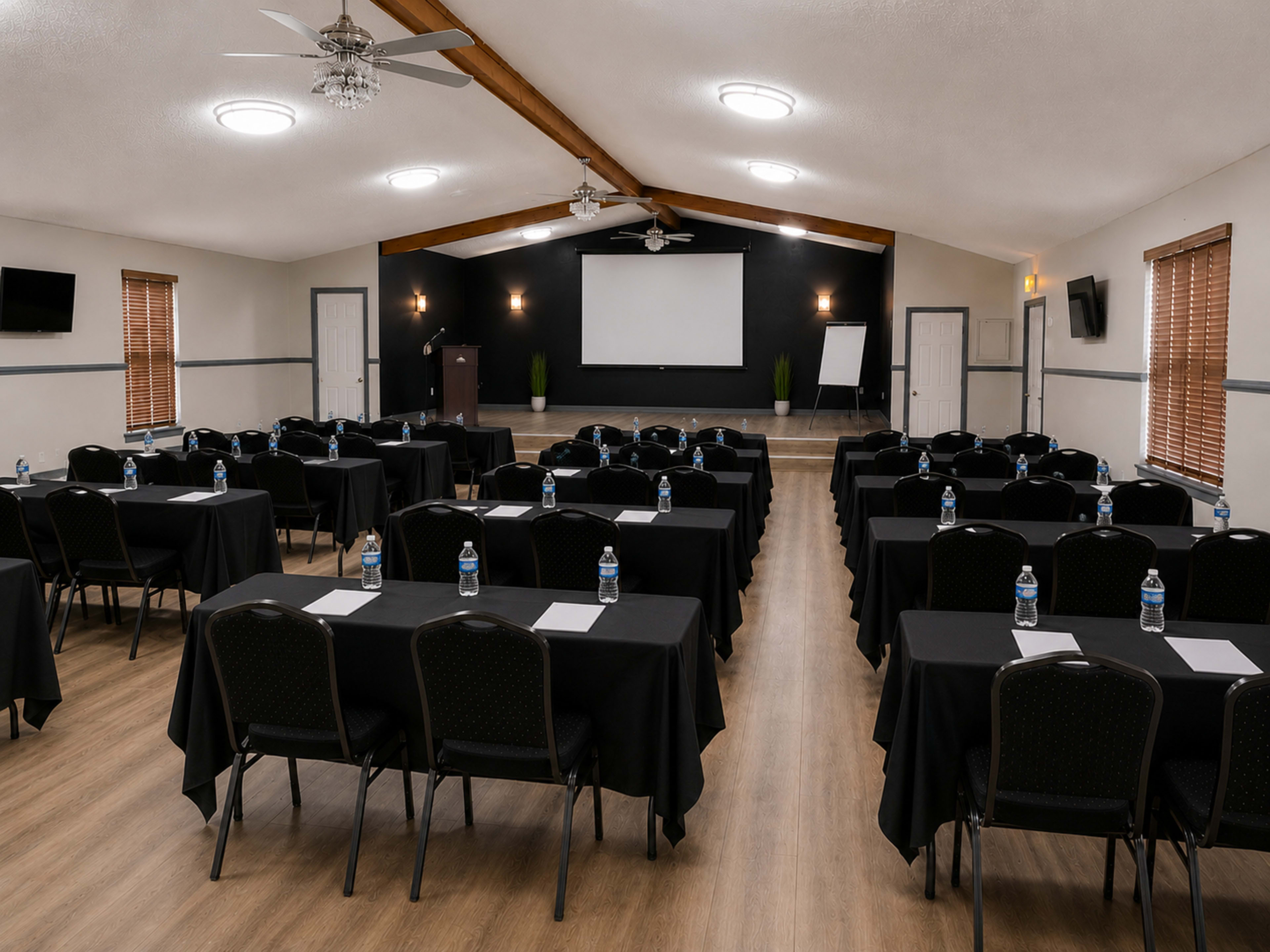 The image shows a conference room set up with rows of black tables and chairs, each adorned with a water bottle and notepad, facing a blank presentation screen at the front.