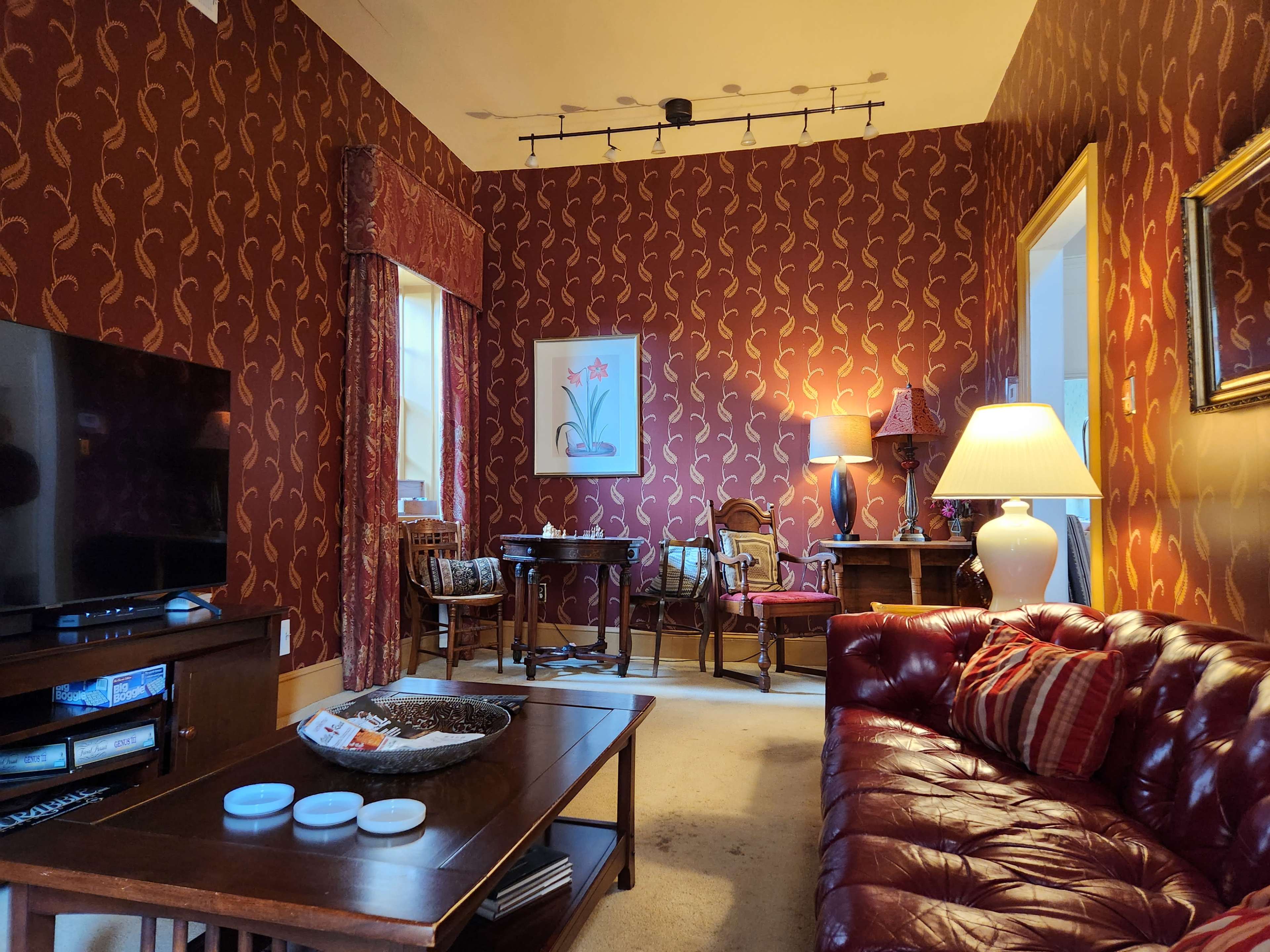 A living room with patterned red wallpaper, a brown leather sofa, a coffee table, a television, and various pieces of furniture, including a lamp and artwork on the walls.