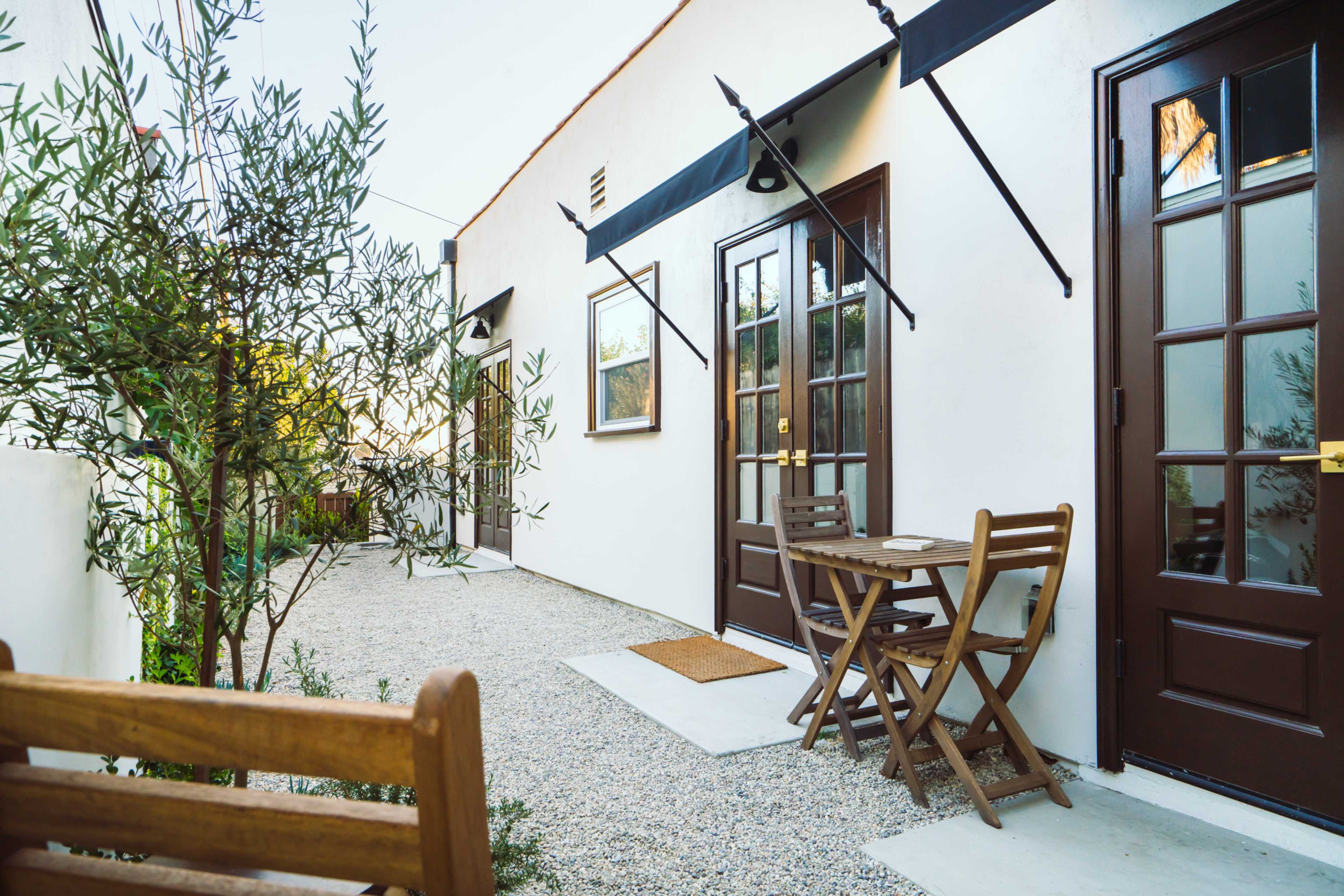 The image shows a narrow pathway lined with olive trees, leading to several doors with dark wooden frames and a small outdoor table with chairs.