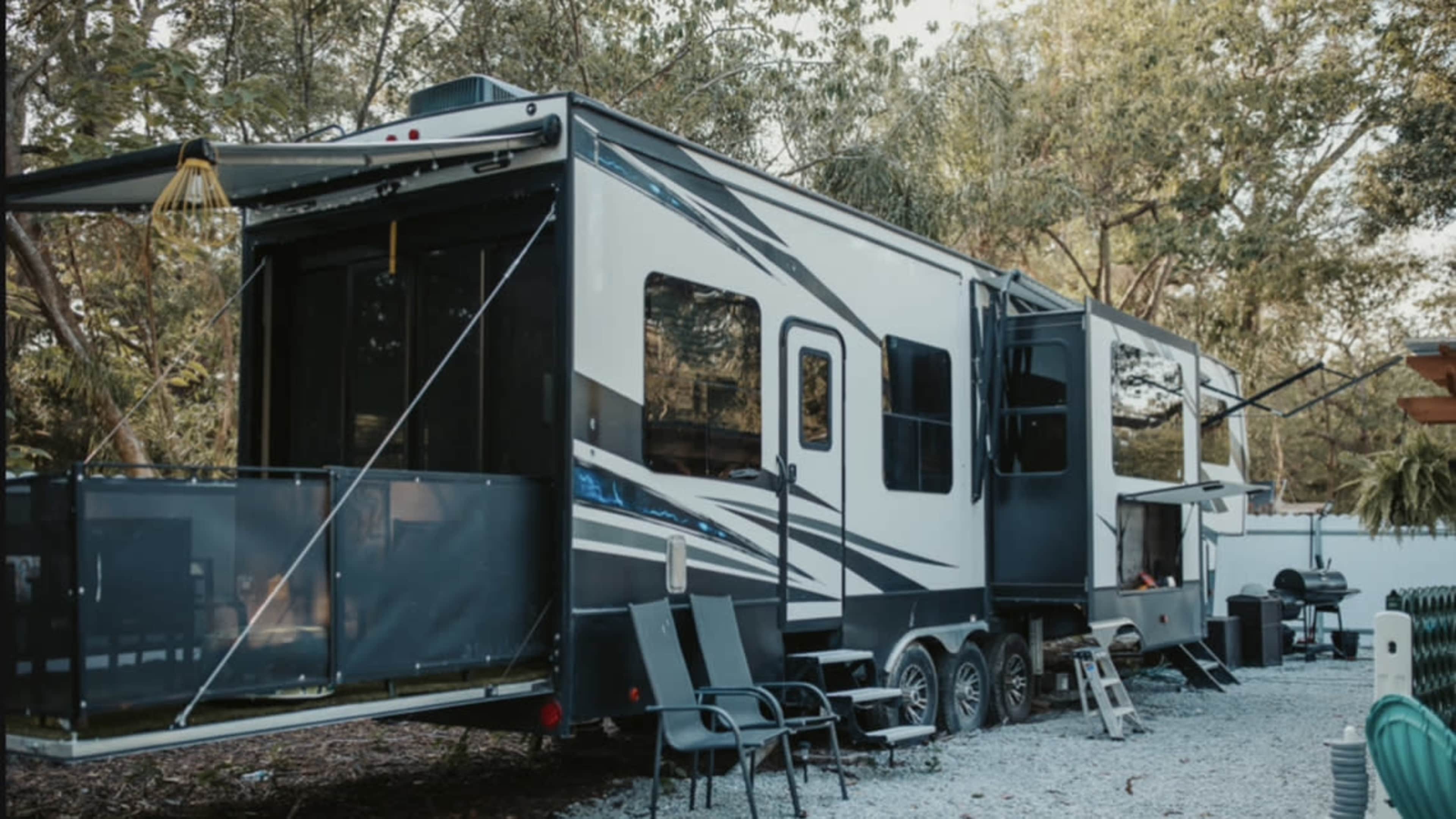 A large, parked RV is situated in a wooded area, surrounded by gravel and equipped with outdoor seating.
