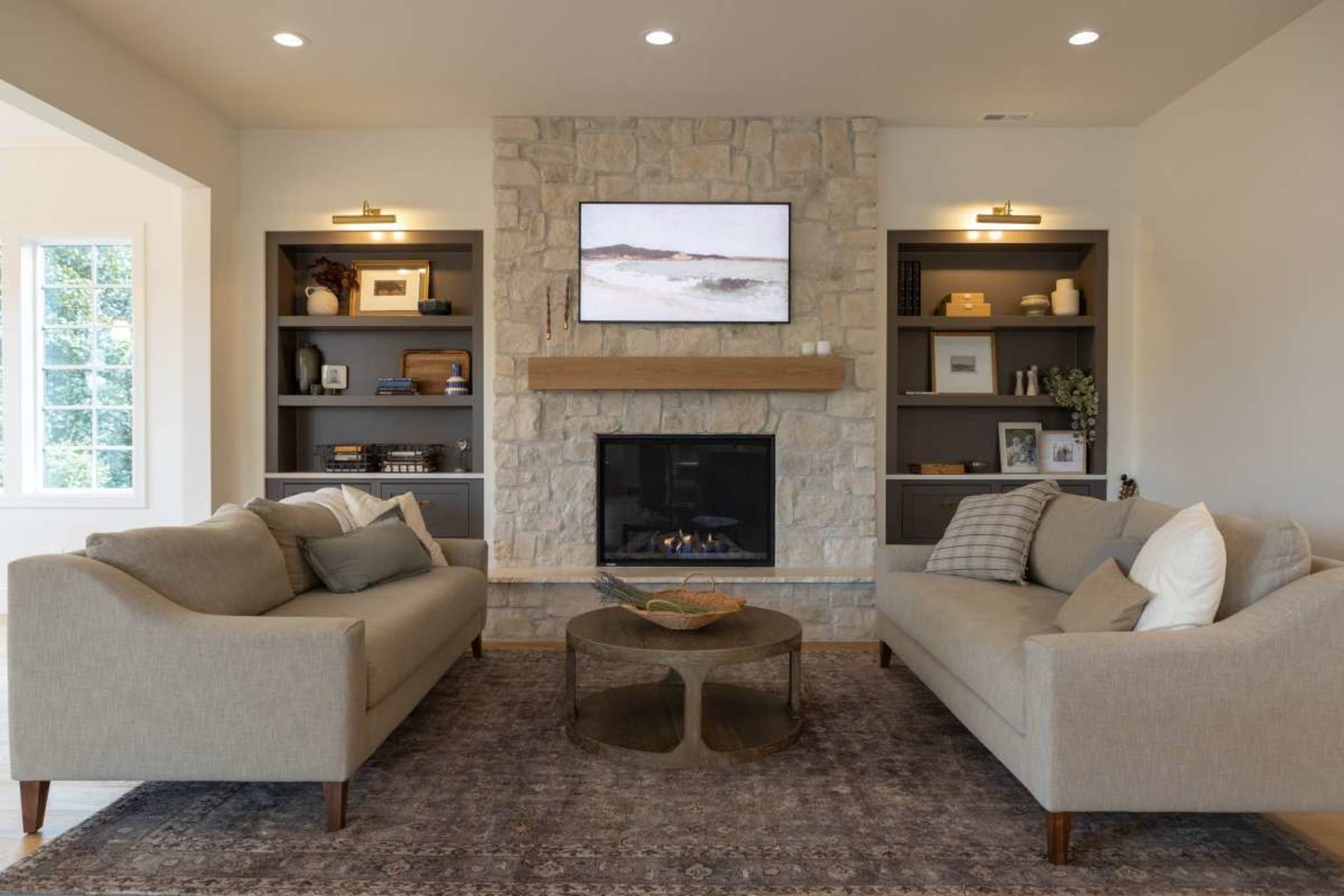 The living room features two beige sofas facing a stone fireplace with a mounted television above, flanked by built-in shelves.