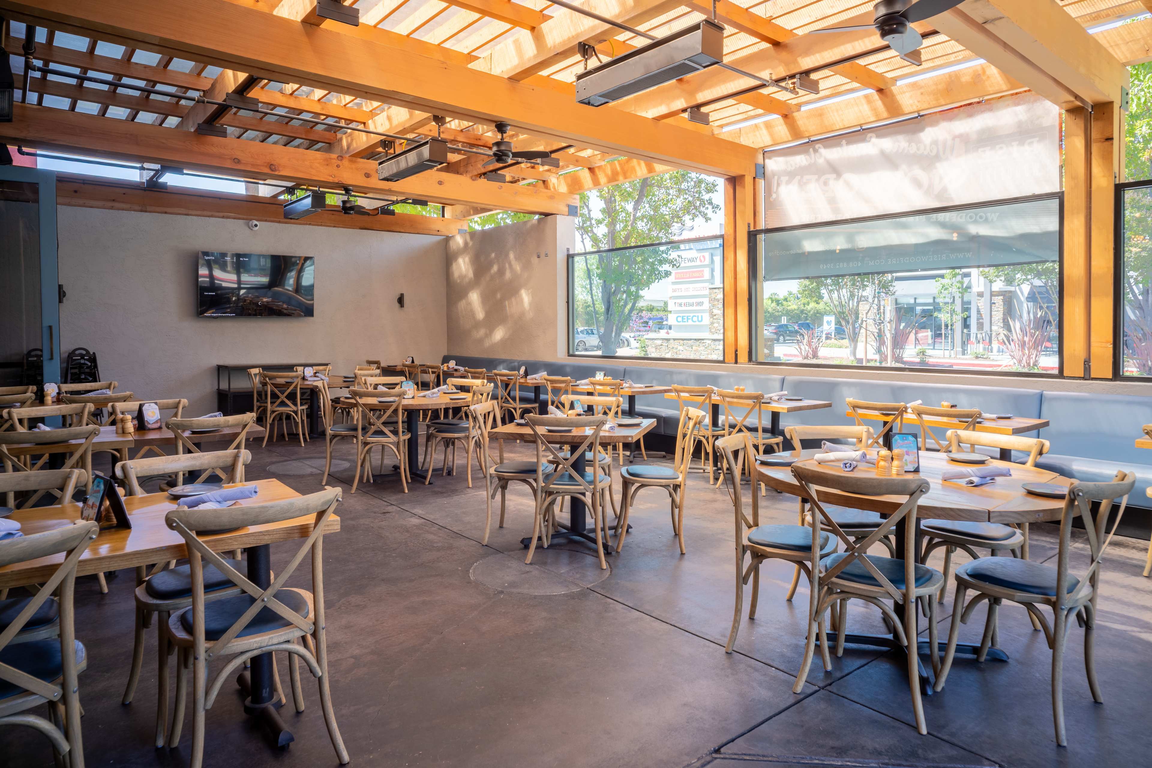 A spacious restaurant interior features wooden tables and chairs arranged neatly under a slatted roof with large windows.