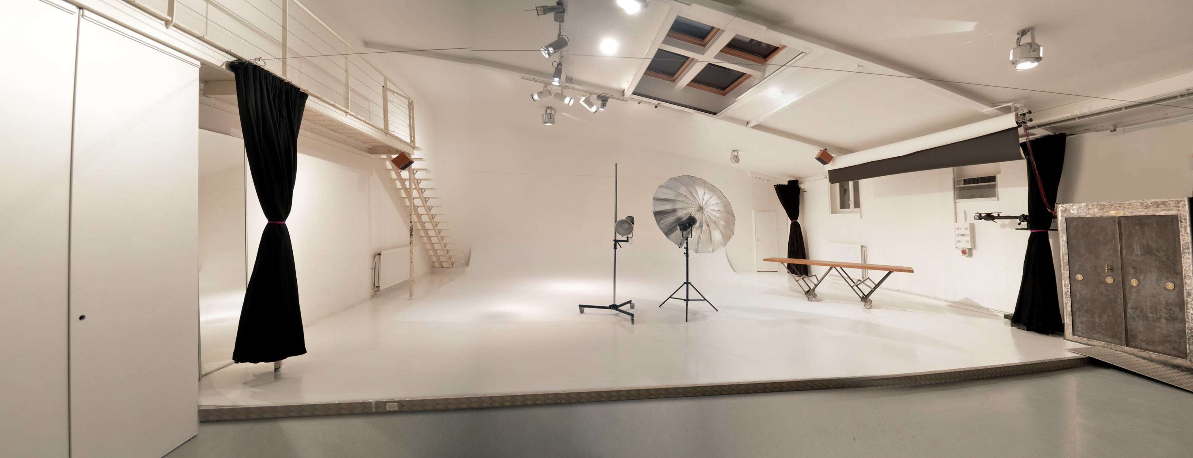 The image shows a spacious, minimalist photography studio with white walls, a flat floor, a large reflector, and a bench.