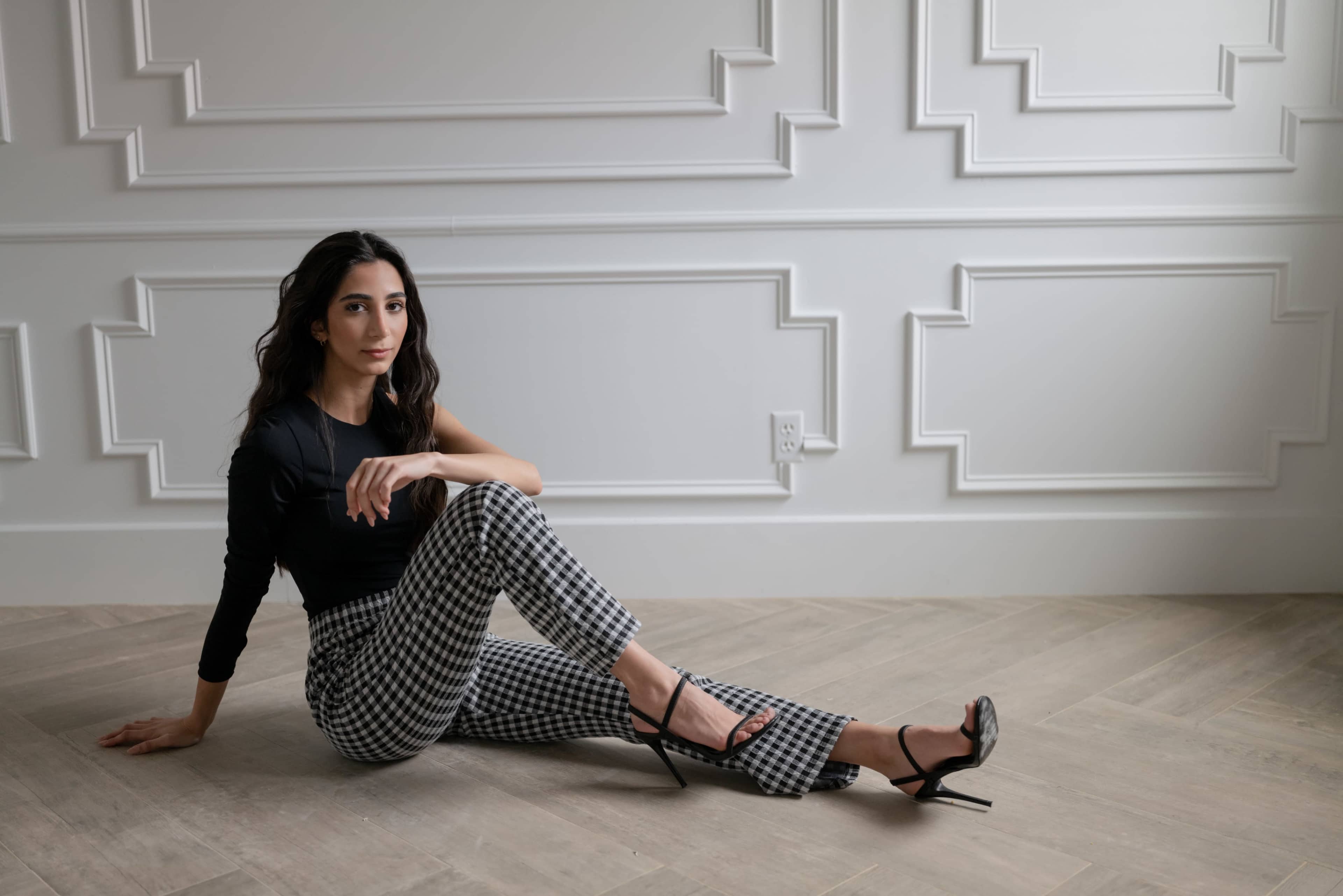 A woman in a black top and checkered pants sits on a light wooden floor against a wall with decorative paneling.