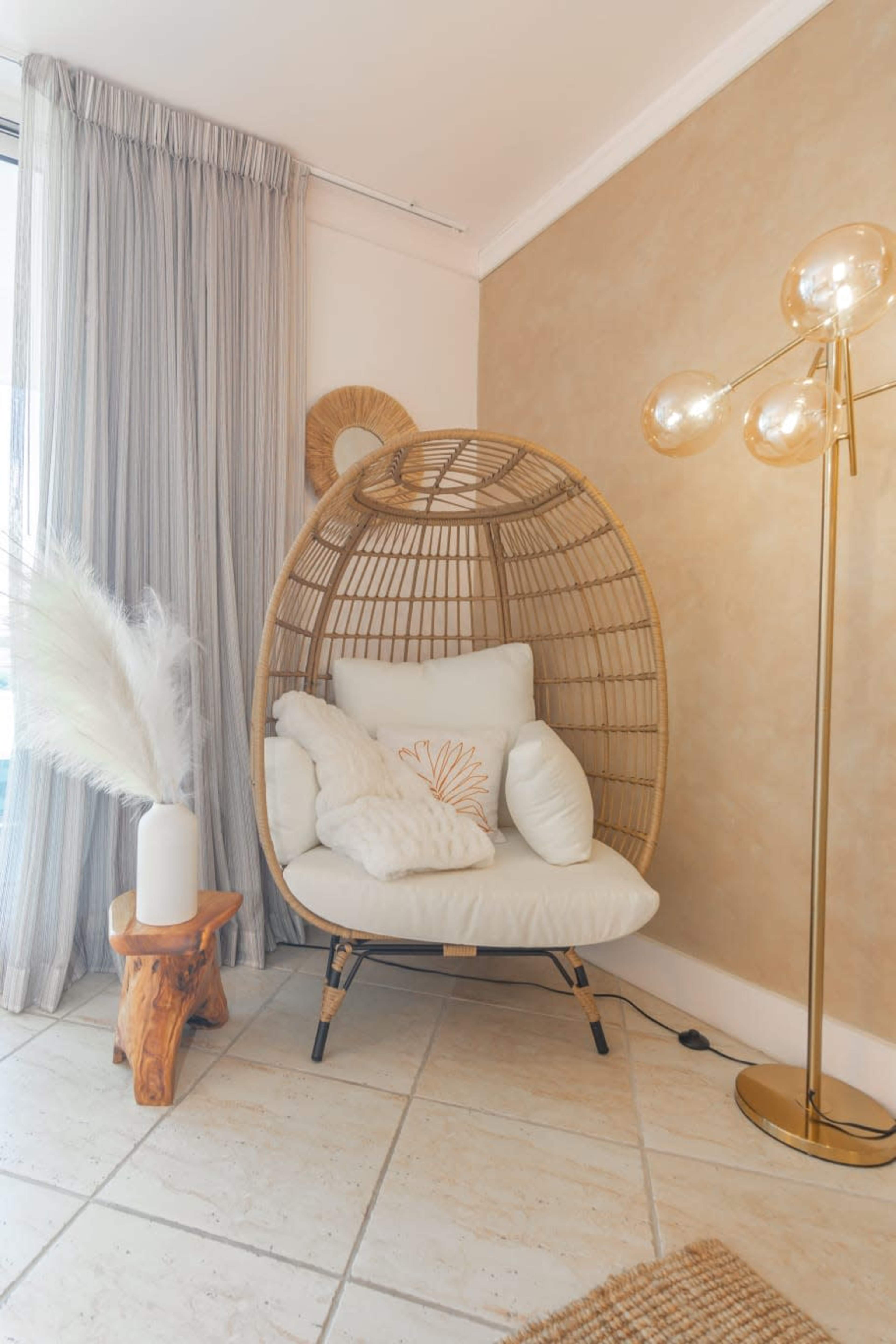 A cozy corner with a rattan lounge chair, a decorative lamp, a small wooden side table, and a vase with dried plants against a textured wall.