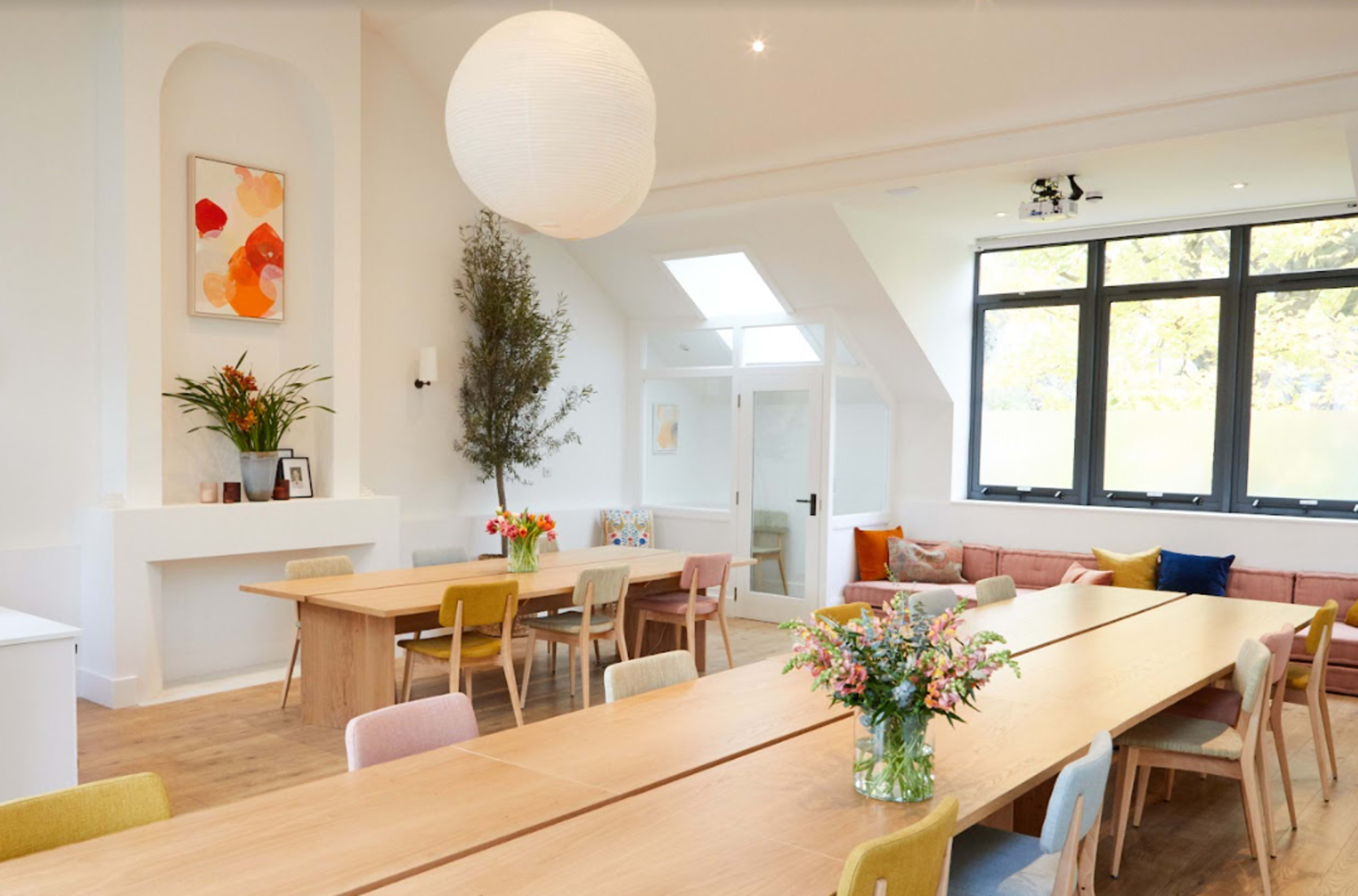 A bright, spacious room with long wooden tables, colorful chairs, and large windows allowing natural light to illuminate the space.