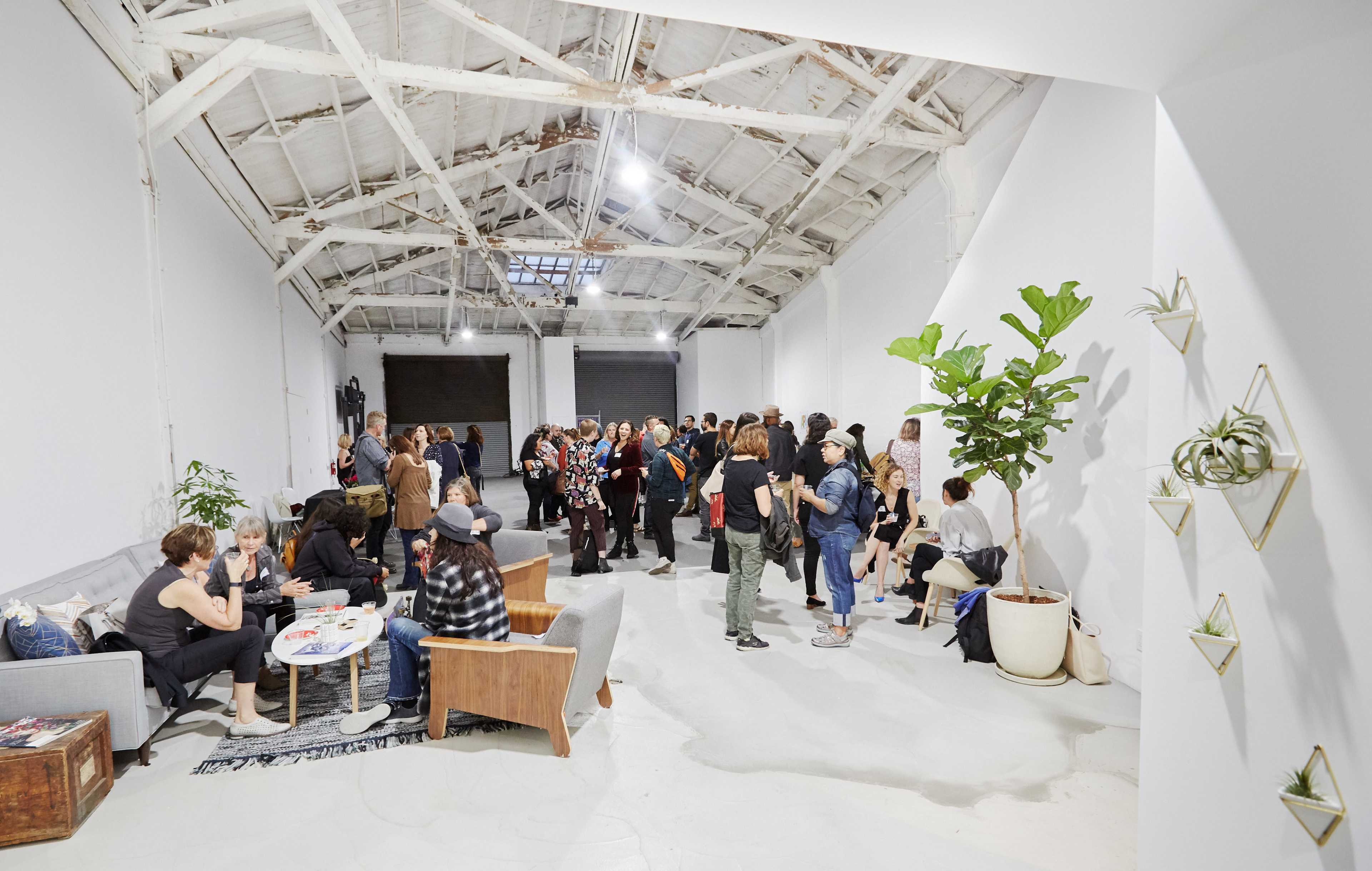 A group of people gather in a spacious, industrial-style room with exposed beams and large windows, socializing and engaging in conversation.