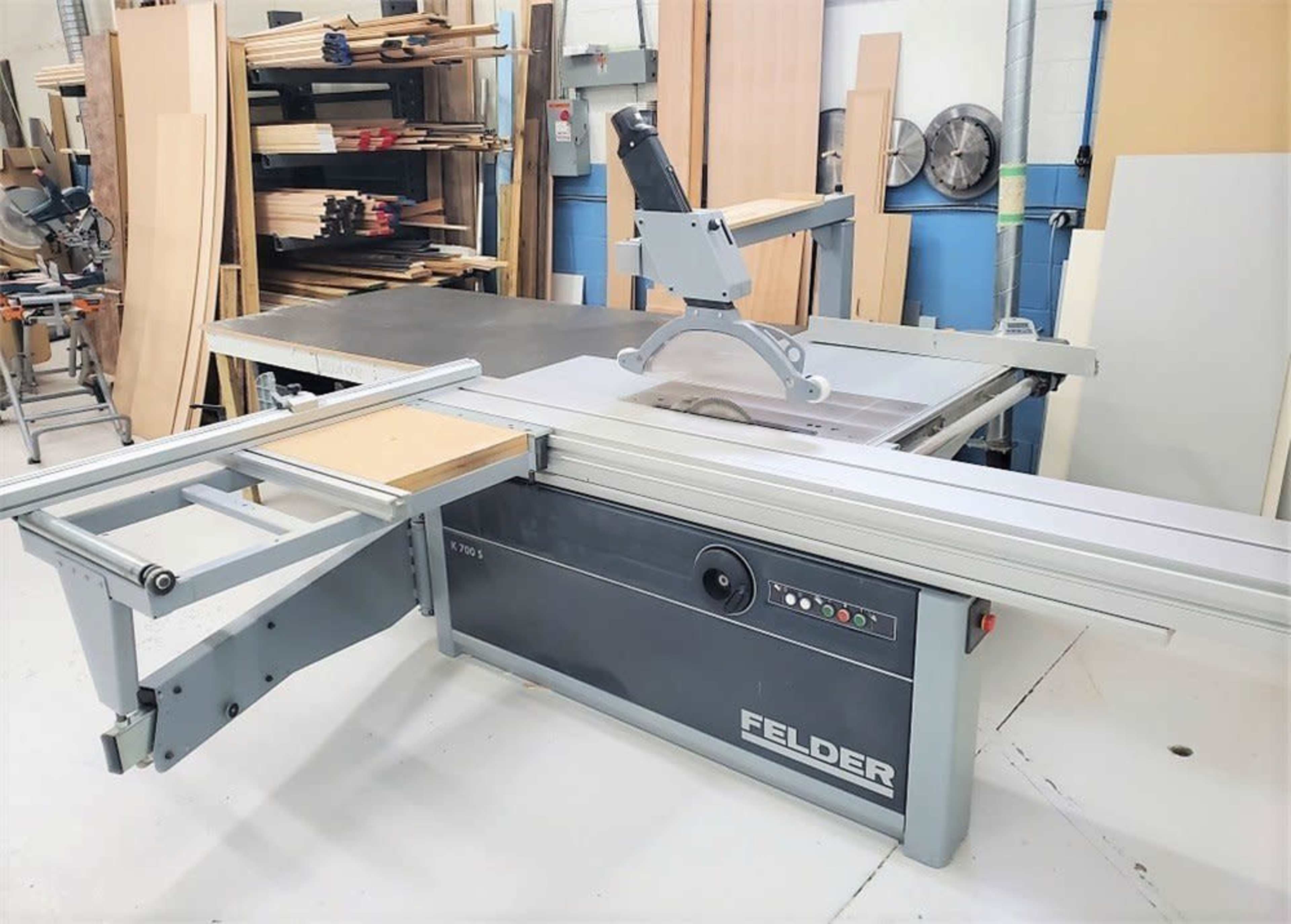 A Felder woodworking machine with a large tabletop and a sliding table saw setup in a workshop surrounded by stacked wood materials.
