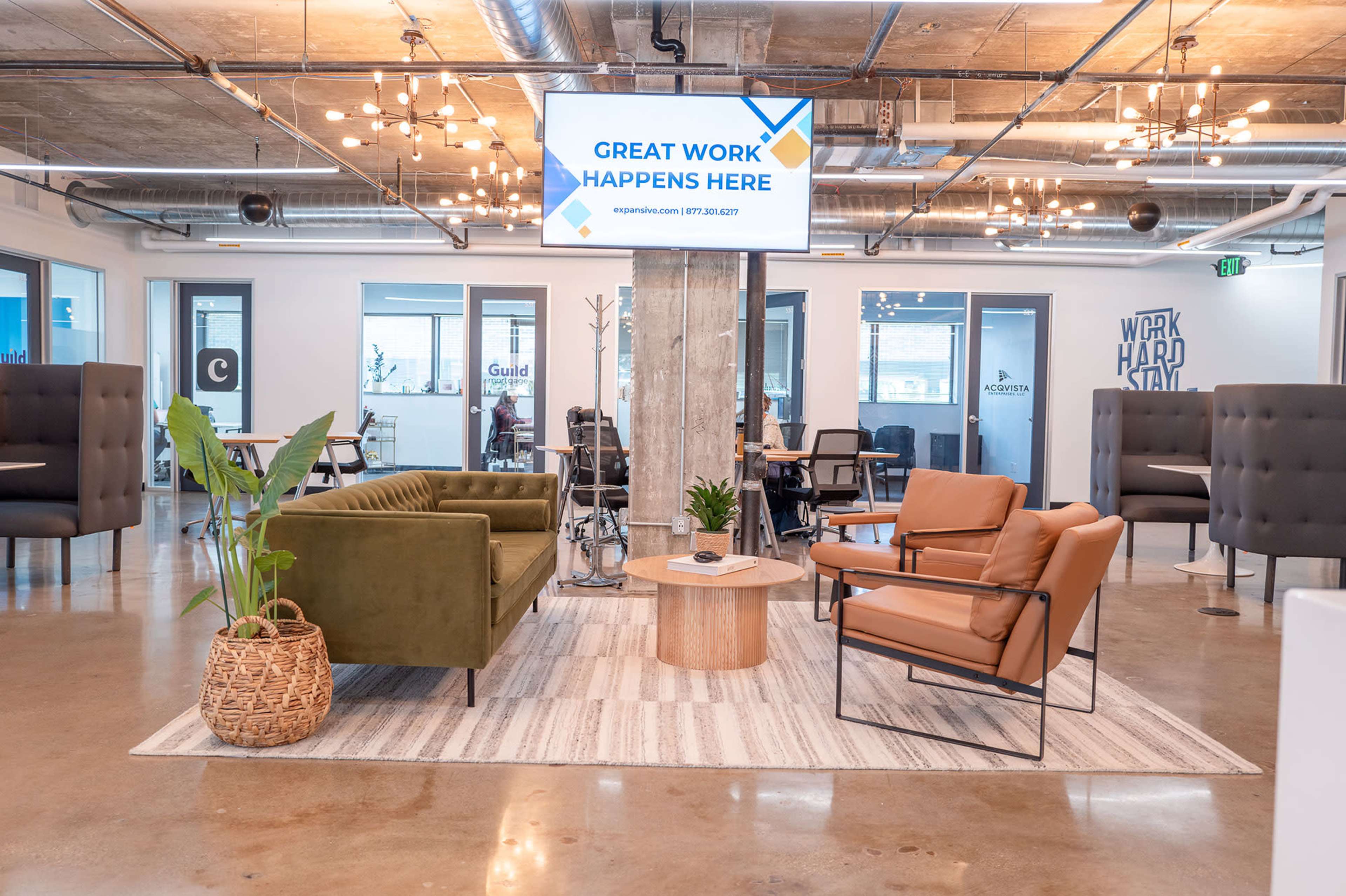 The image shows a modern office lounge area with comfortable seating, a coffee table, and a large screen displaying a sign that reads "Great Work Happens Here."