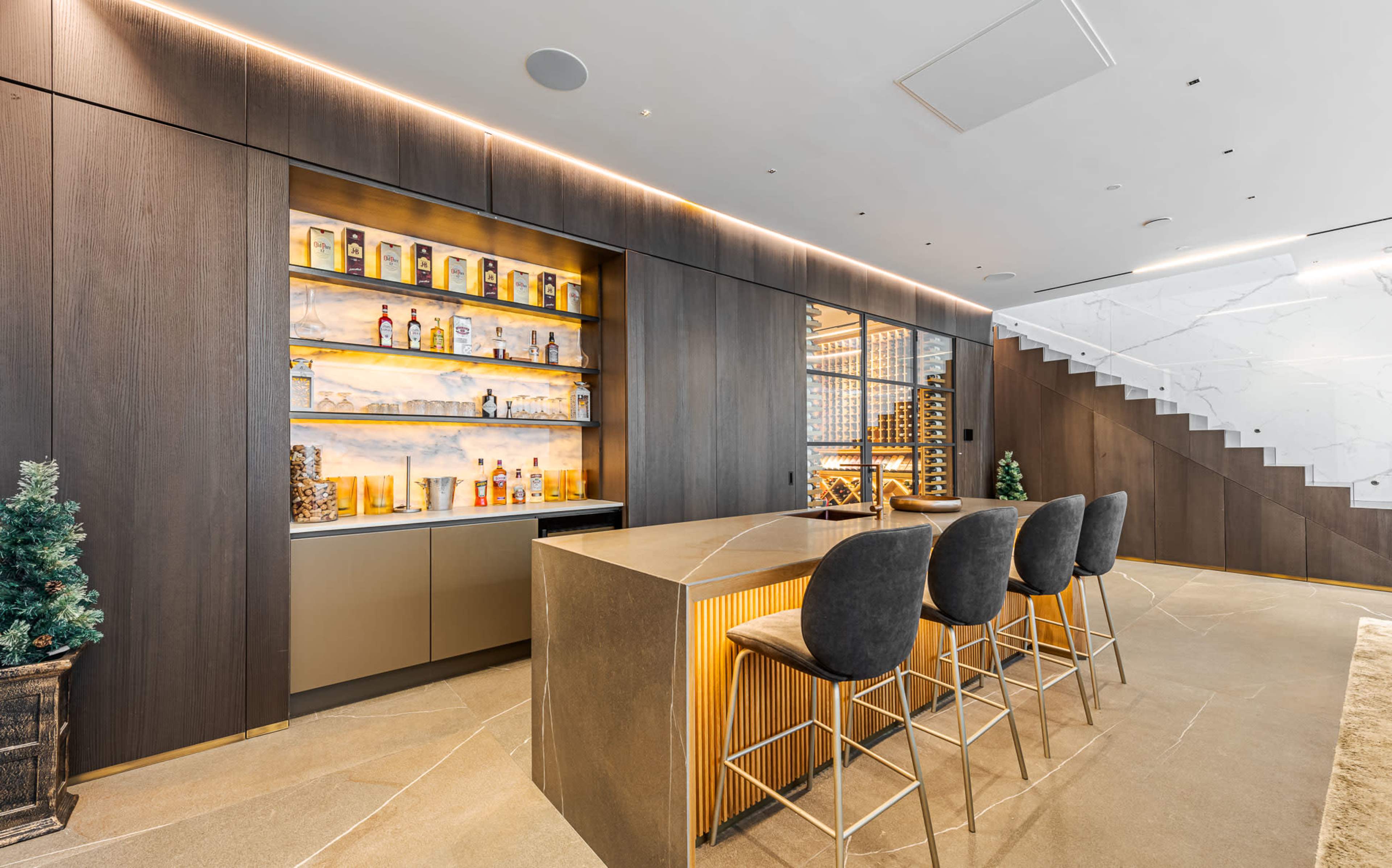 A modern home bar features a backlit shelf displaying various bottles and four bar stools in front of a smooth countertop.