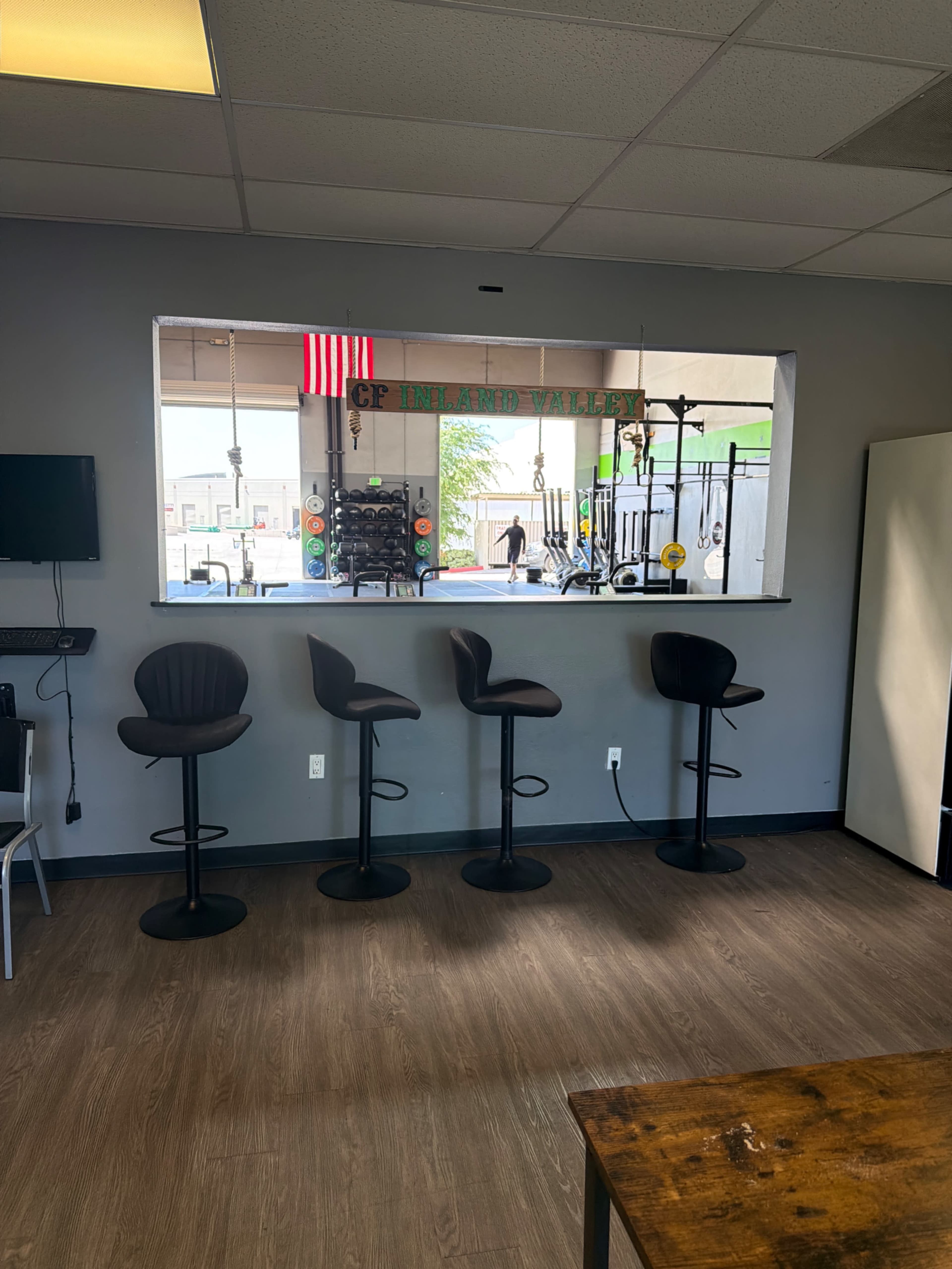 The image shows a view from an indoor space with four modern bar stools arranged in front of a window, through which gym equipment and an American flag are visible outside.