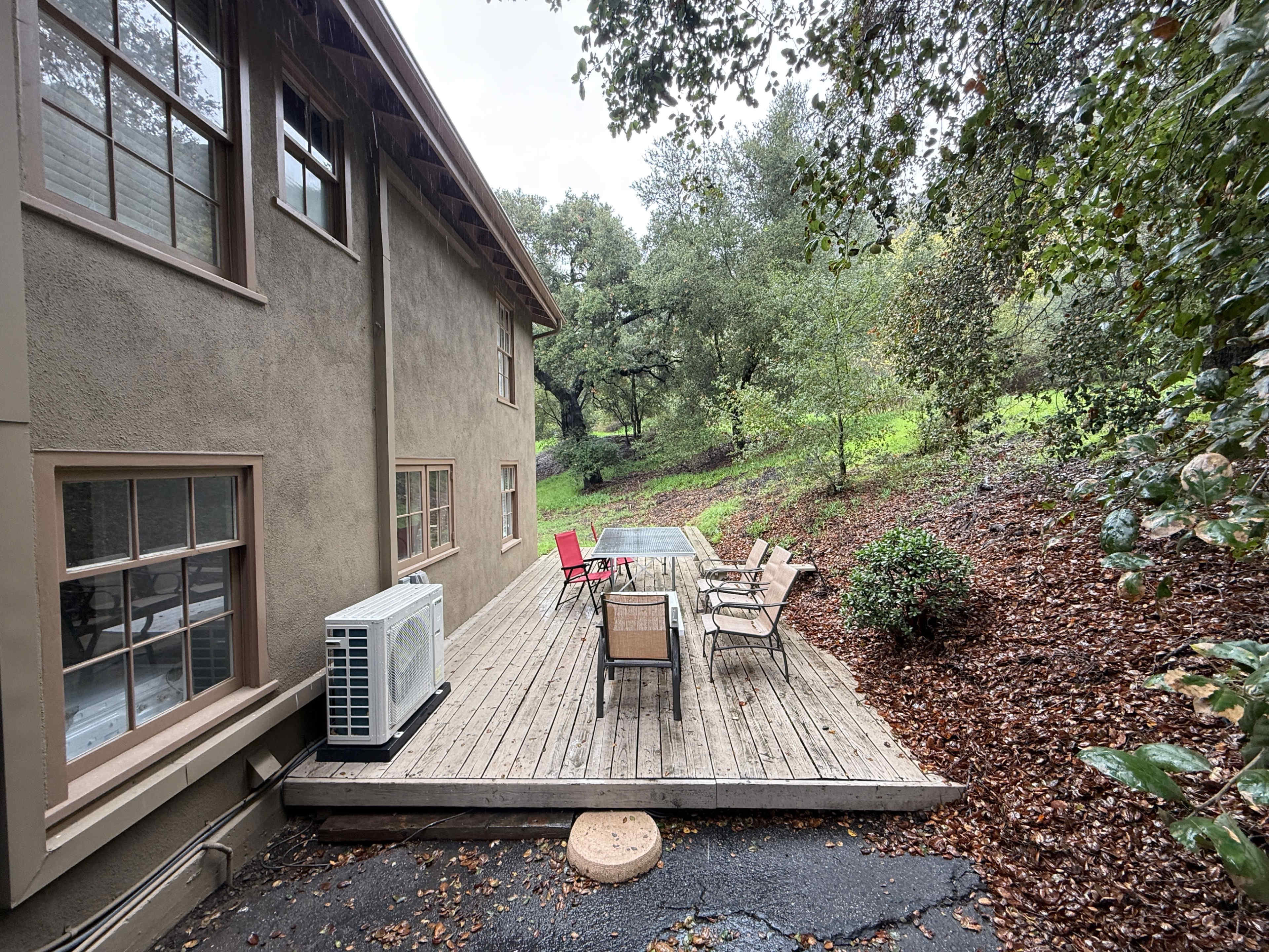 A wooden deck with several chairs and a table is attached to the side of a house, surrounded by trees and fallen leaves.