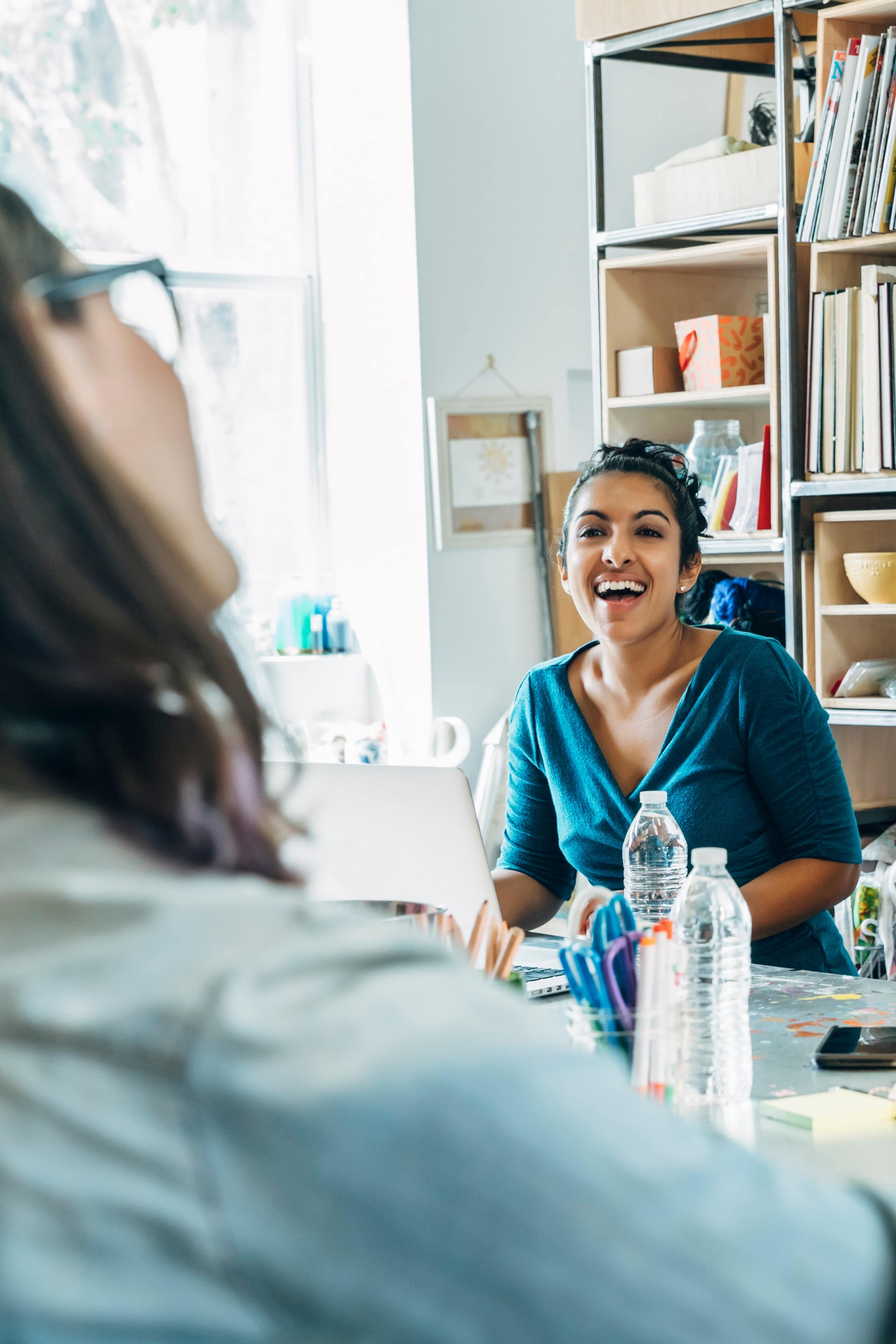 A woman in a blue shirt smiles while sitting at a desk with a laptop and bottles of water in a bright room filled with books and art supplies.