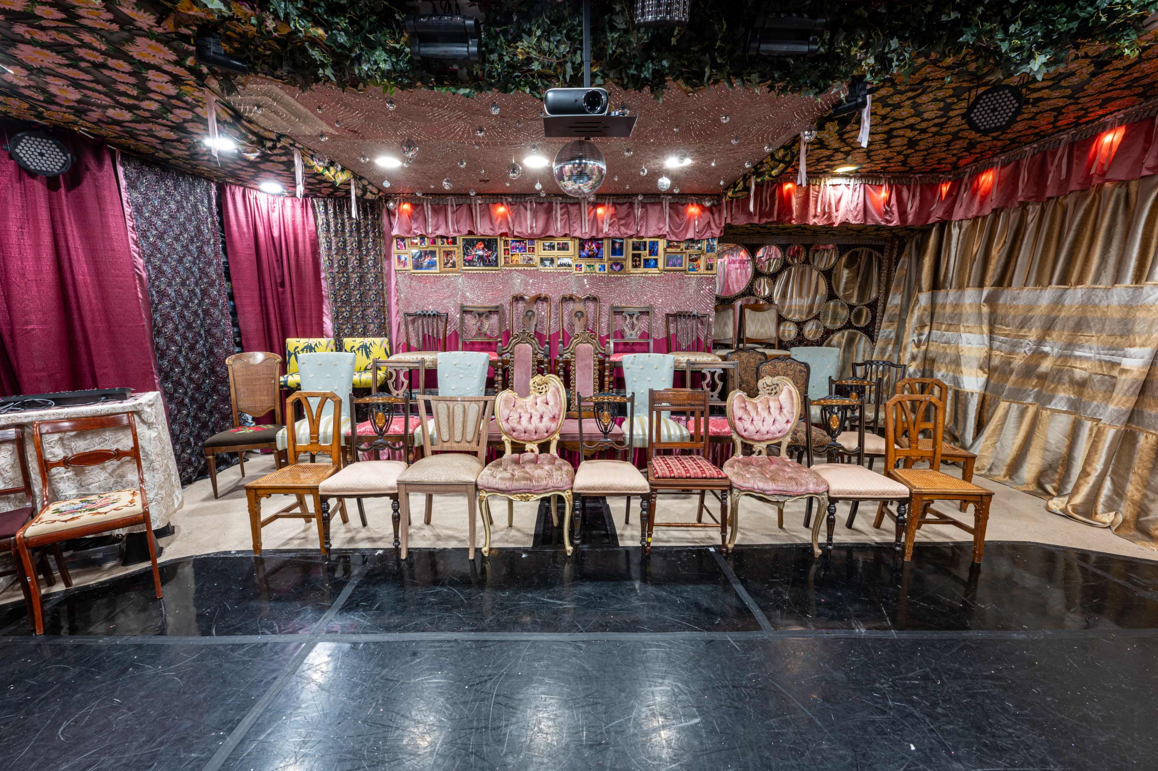 The image shows a decorated event space with a row of various chairs arranged in front of a backdrop featuring colorful drapes and framed photographs.