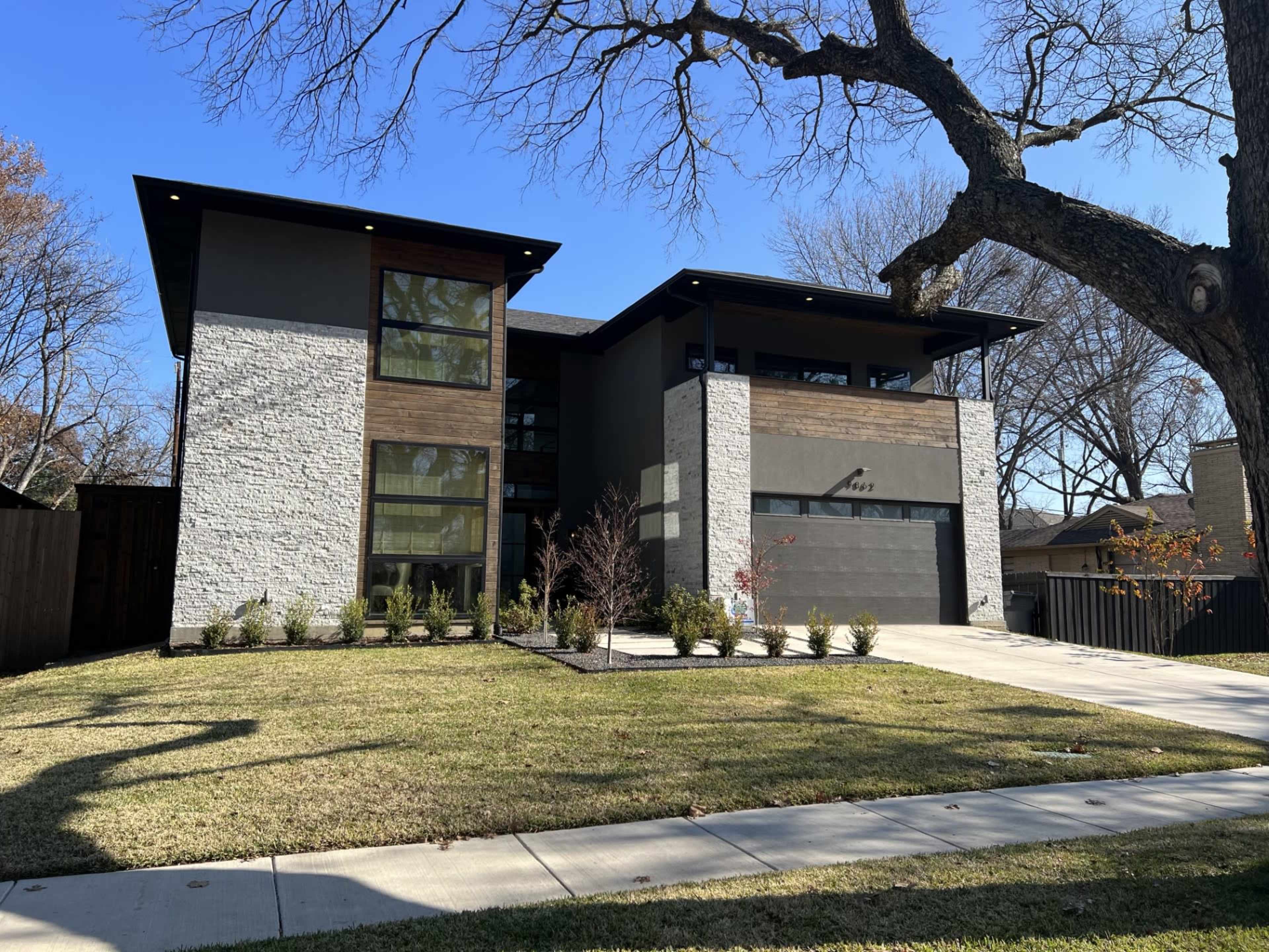 A modern two-story house features a mix of stone and wood exterior, with large windows and a landscaped front yard.