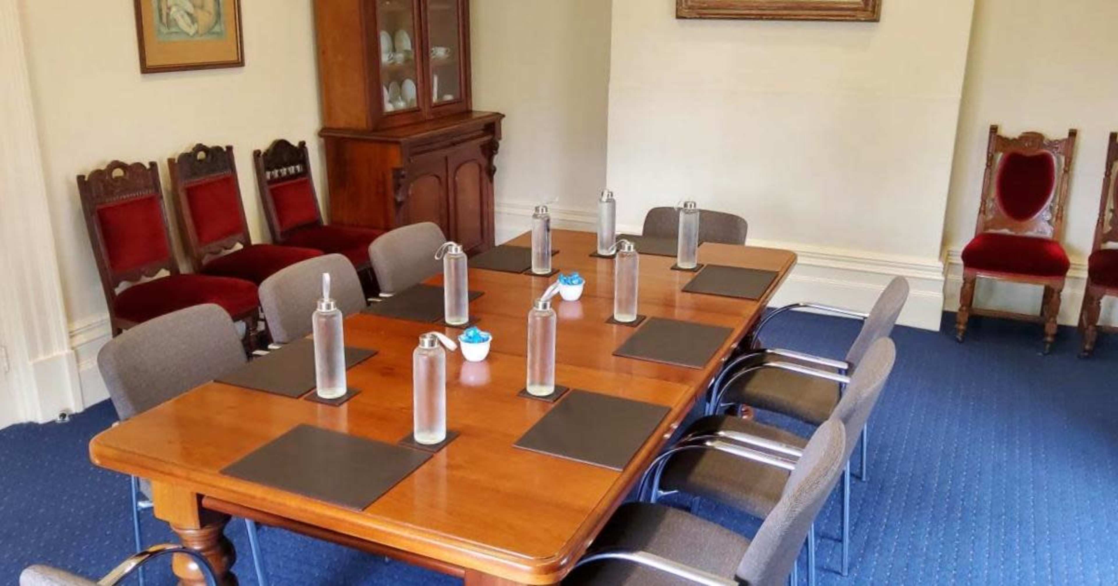 A rectangular wooden table with placemats and water bottles is surrounded by chairs in a light-colored meeting room.
