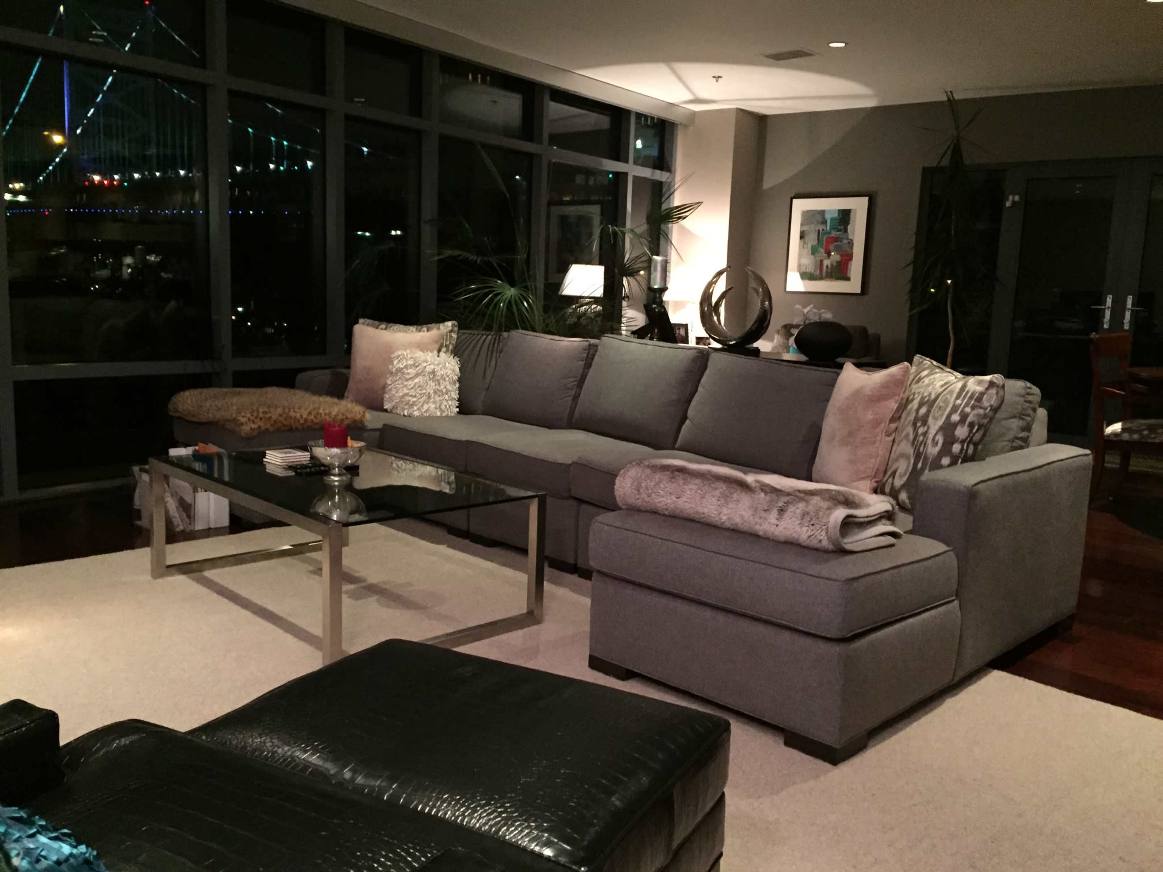 A modern living room features a large gray sectional sofa with decorative pillows, a glass coffee table, and floor-to-ceiling windows showcasing a view of the city skyline at night.