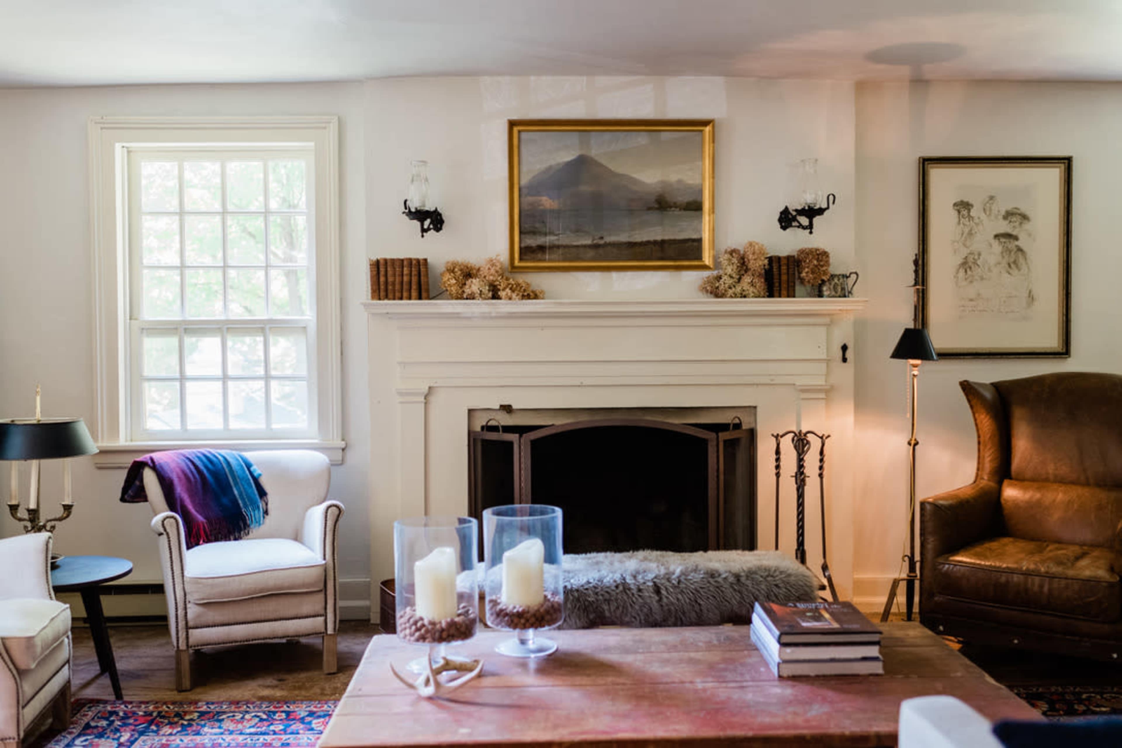 The image shows a cozy living room featuring a fireplace with decorative candles, a comfortable armchair, a sofa with a throw blanket, and a coffee table adorned with books and decorative items.