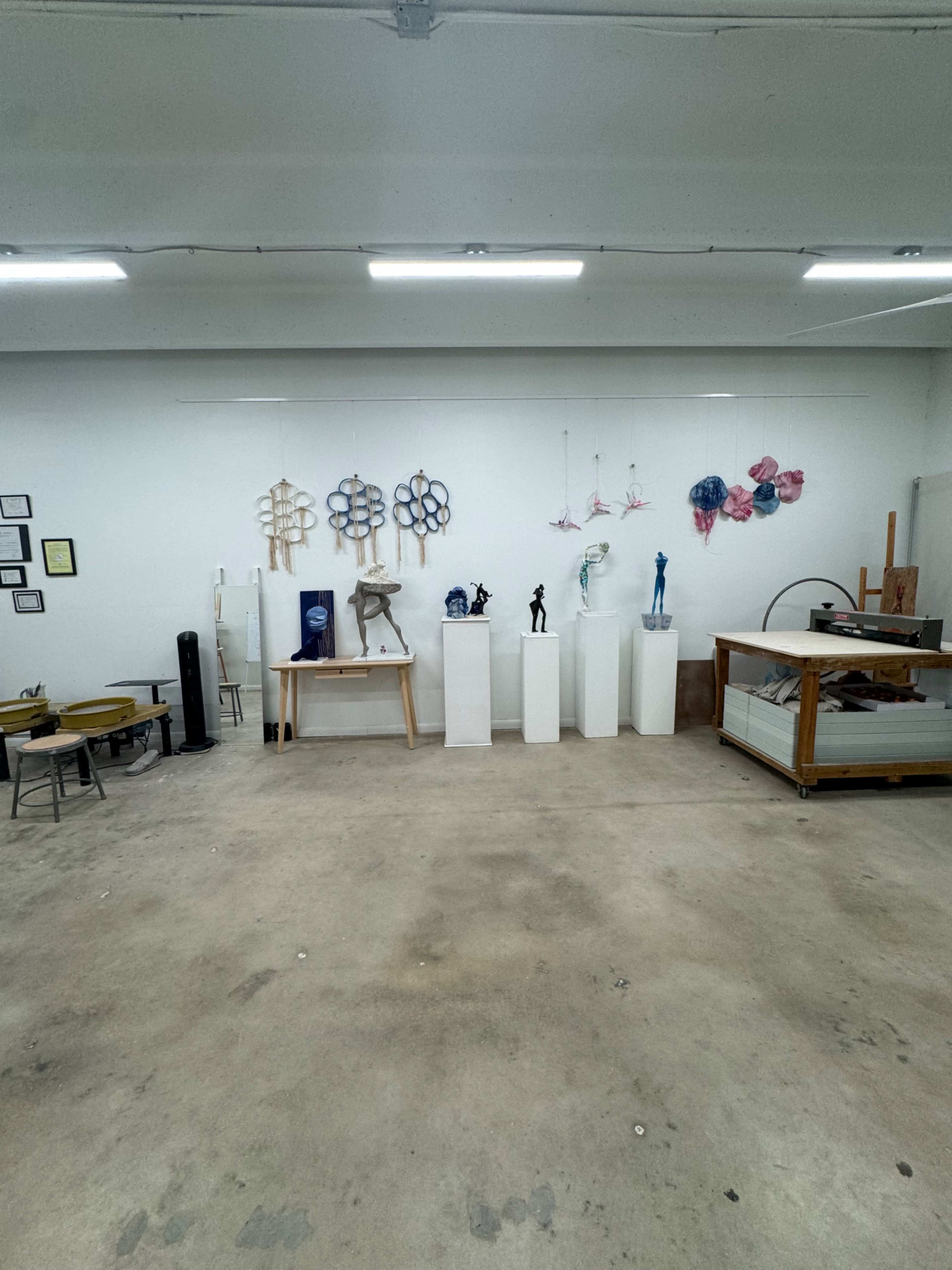 The image shows an art studio with various sculptures displayed on pedestals against a white wall, along with a circular table and tools in the foreground.