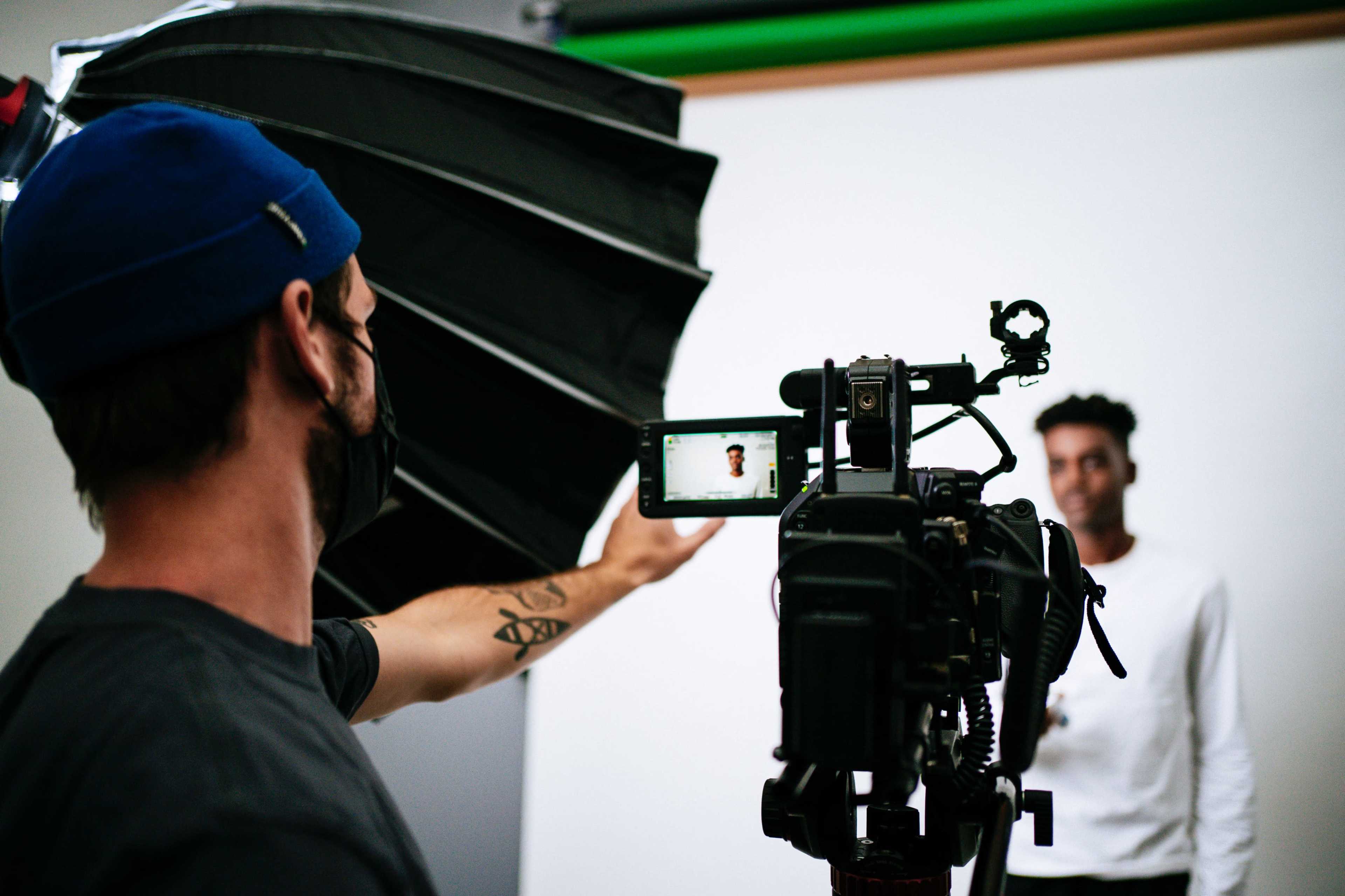 A photographer in a blue cap and mask holds a camera directed at a smiling subject in a white shirt against a plain backdrop.