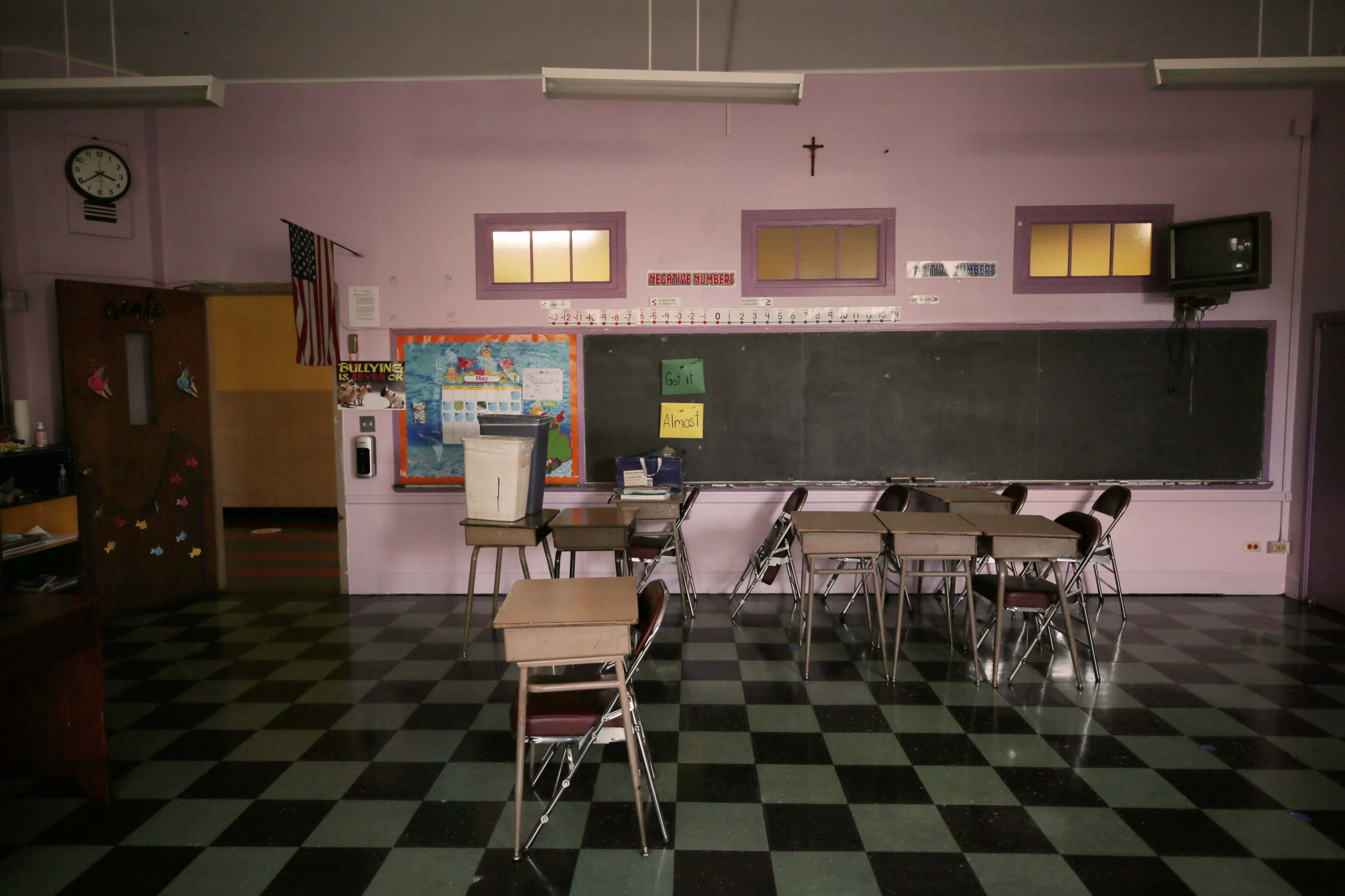 A classroom features empty chairs and desks arranged in groups, with a chalkboard, bulletin board, and a clock on the wall.