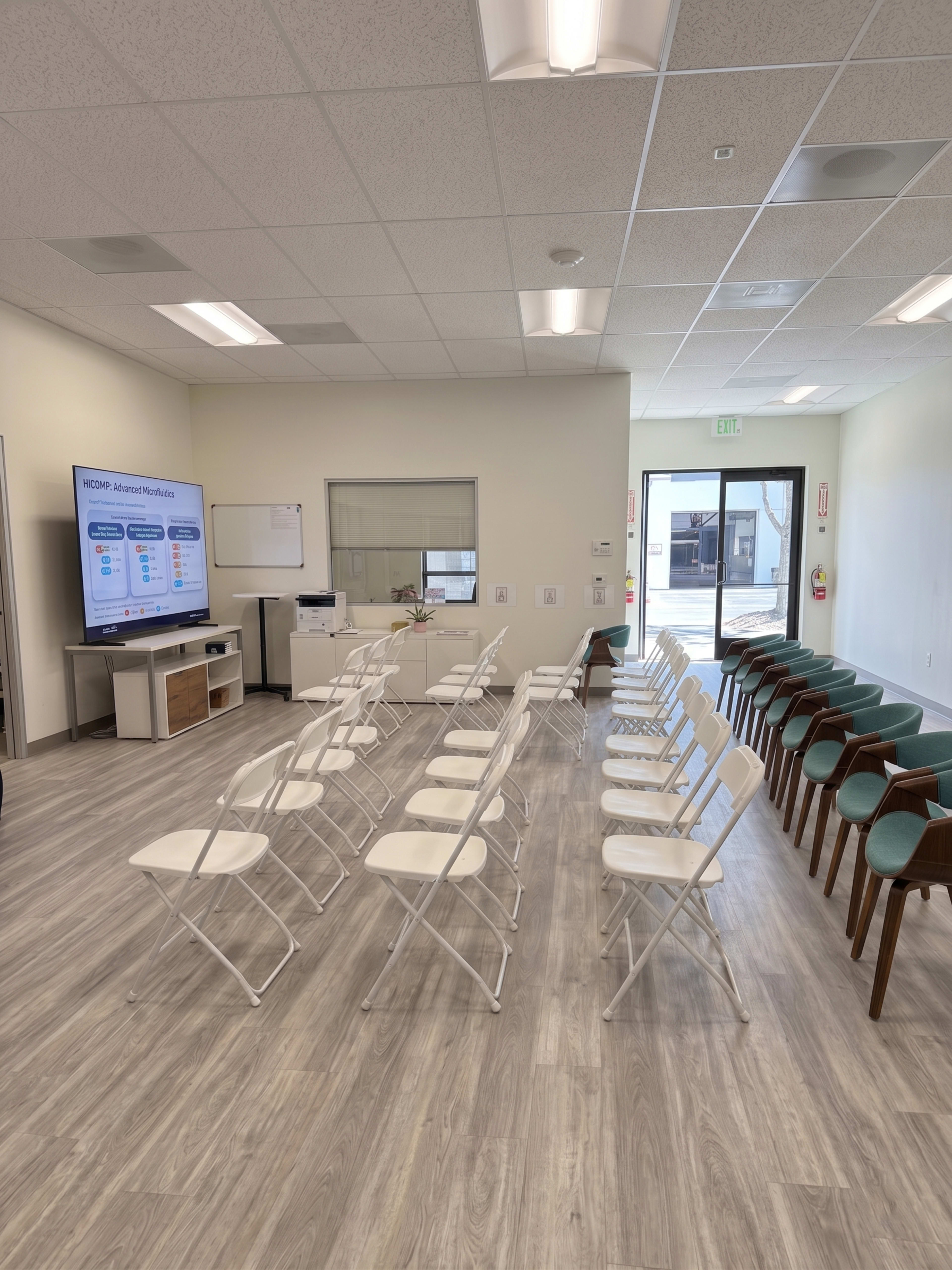 The image shows a waiting room with several rows of white folding chairs arranged in front of a large screen displaying information, and a mix of green and wooden chairs along one side.