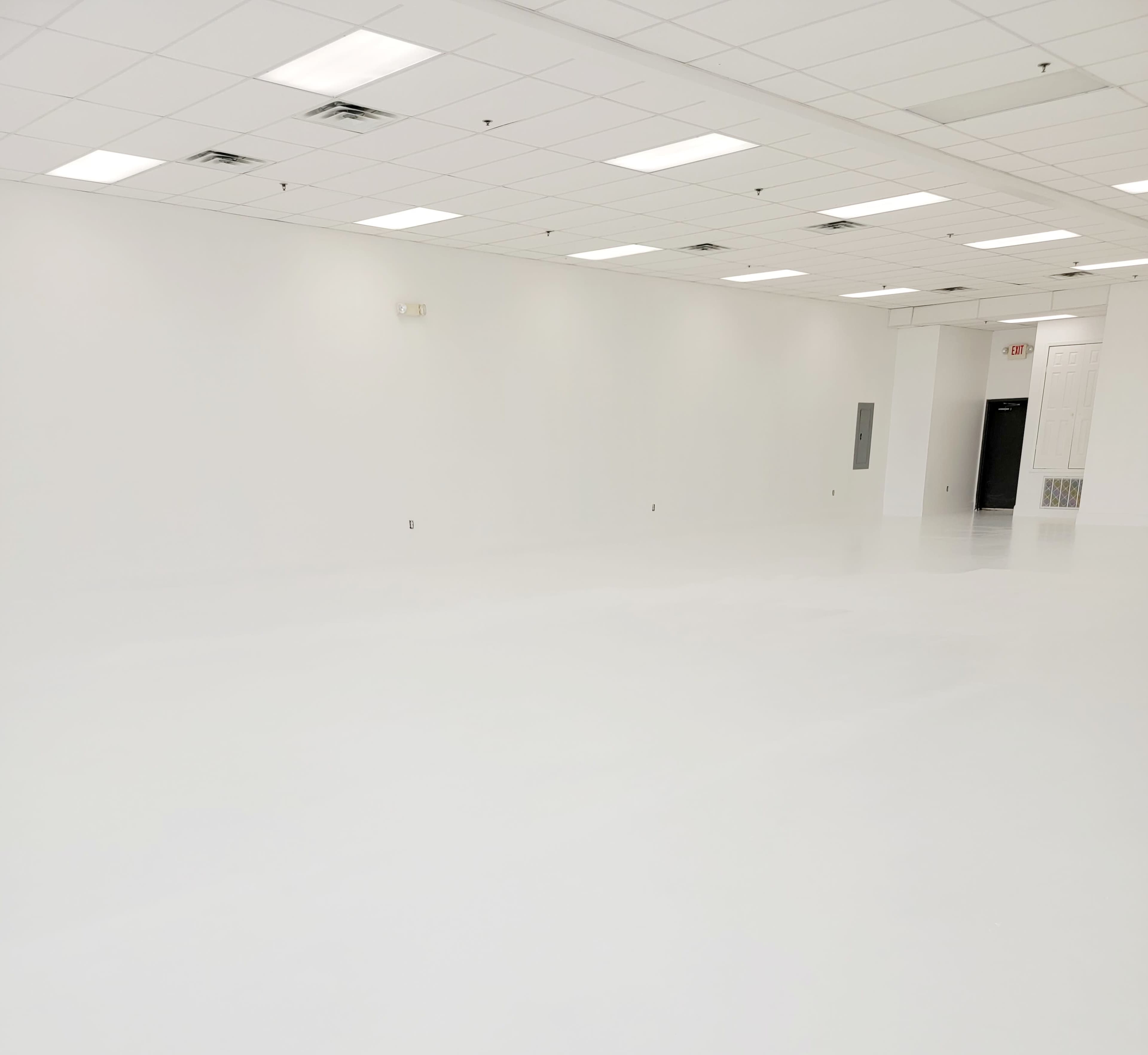 The image shows an empty room with white walls and a white floor, featuring a few ceiling lights and a single door on one side.