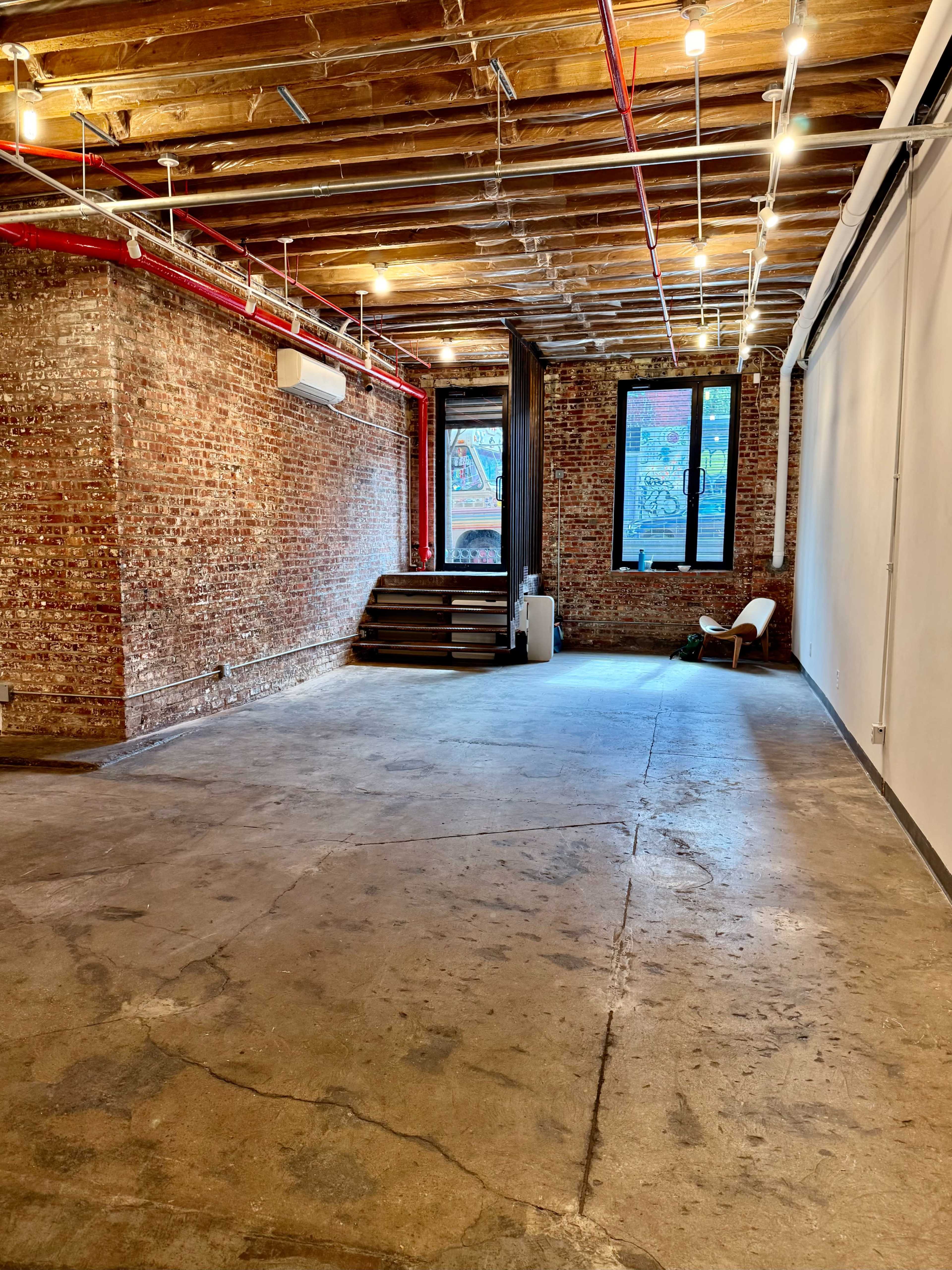 The image shows an empty industrial-style room with exposed brick walls, wooden beams, and a concrete floor, featuring a staircase leading to a window.