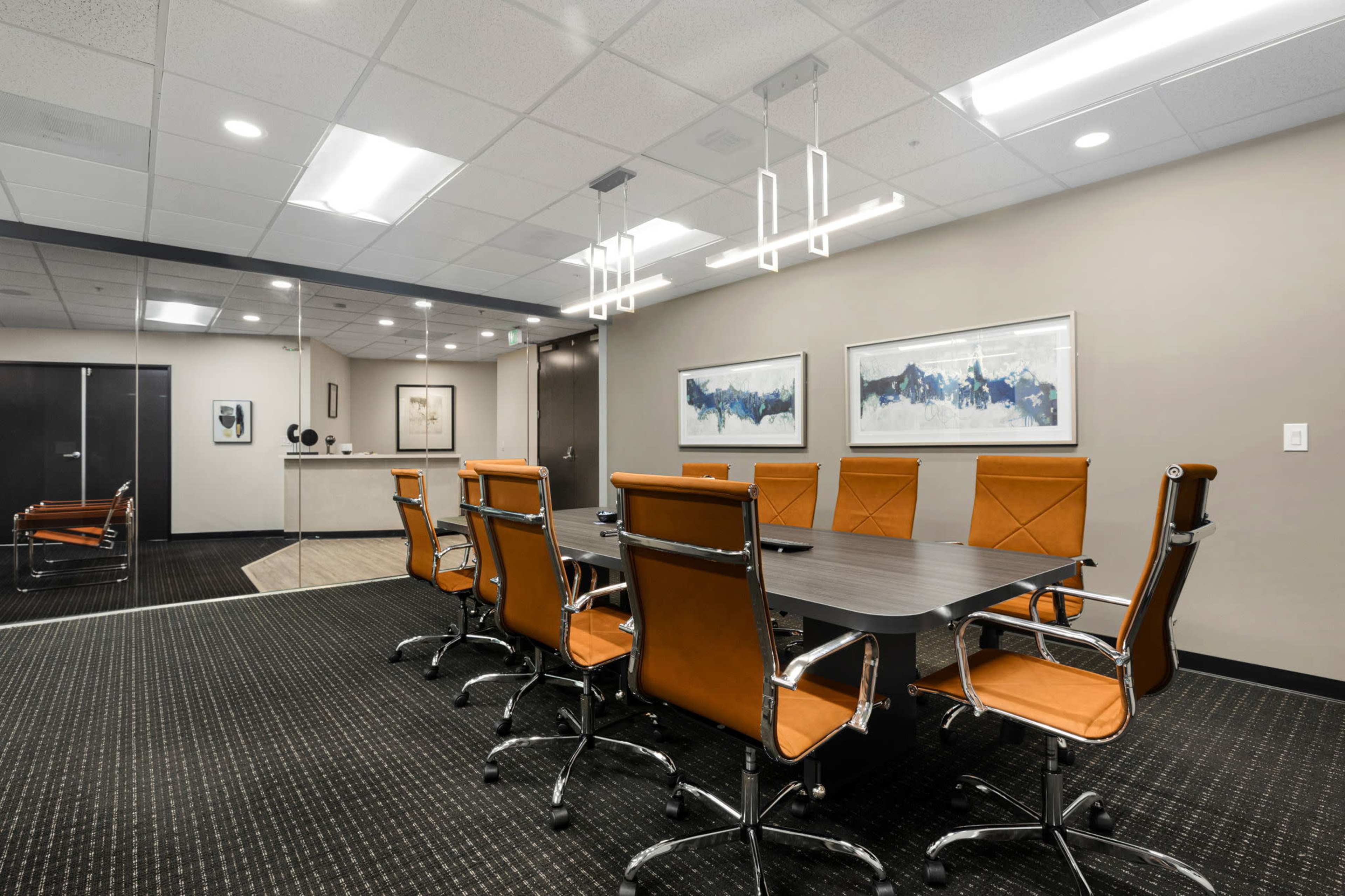 A modern conference room featuring a large rectangular table surrounded by eight orange leather chairs, with a glass wall in the background.