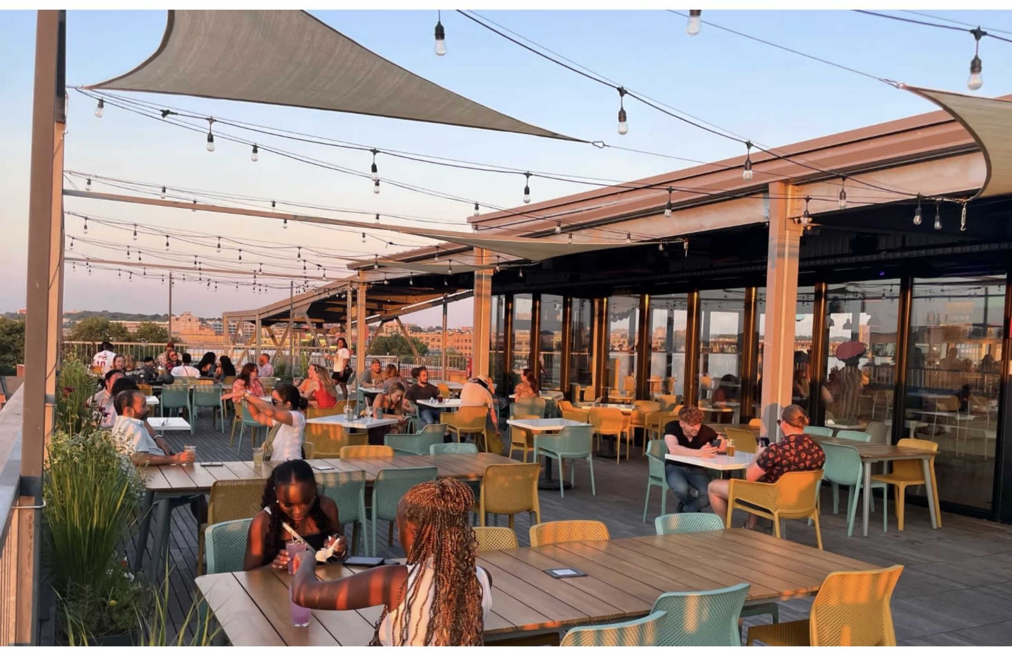 The image shows a rooftop dining area filled with people seated at tables, surrounded by decorative string lights and shaded by awnings.