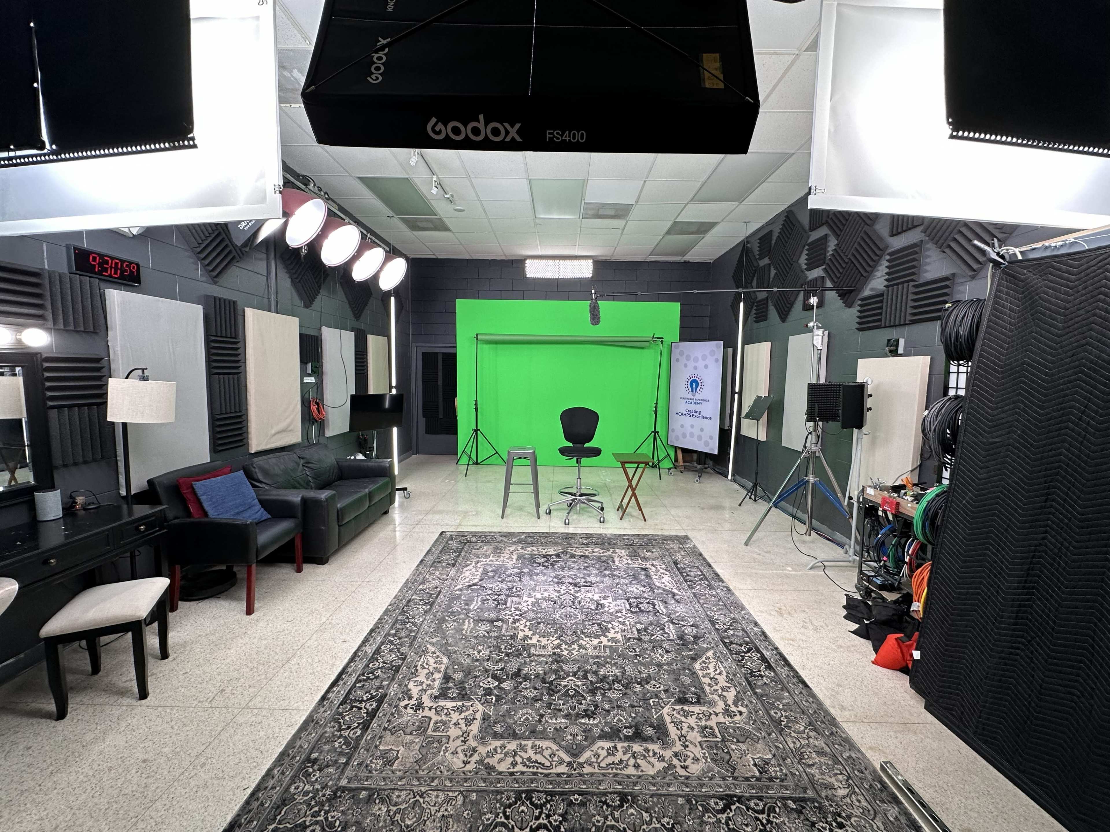 The image shows a well-lit studio space with a green screen at one end, surrounded by various equipment, seating areas, and soundproofed walls.