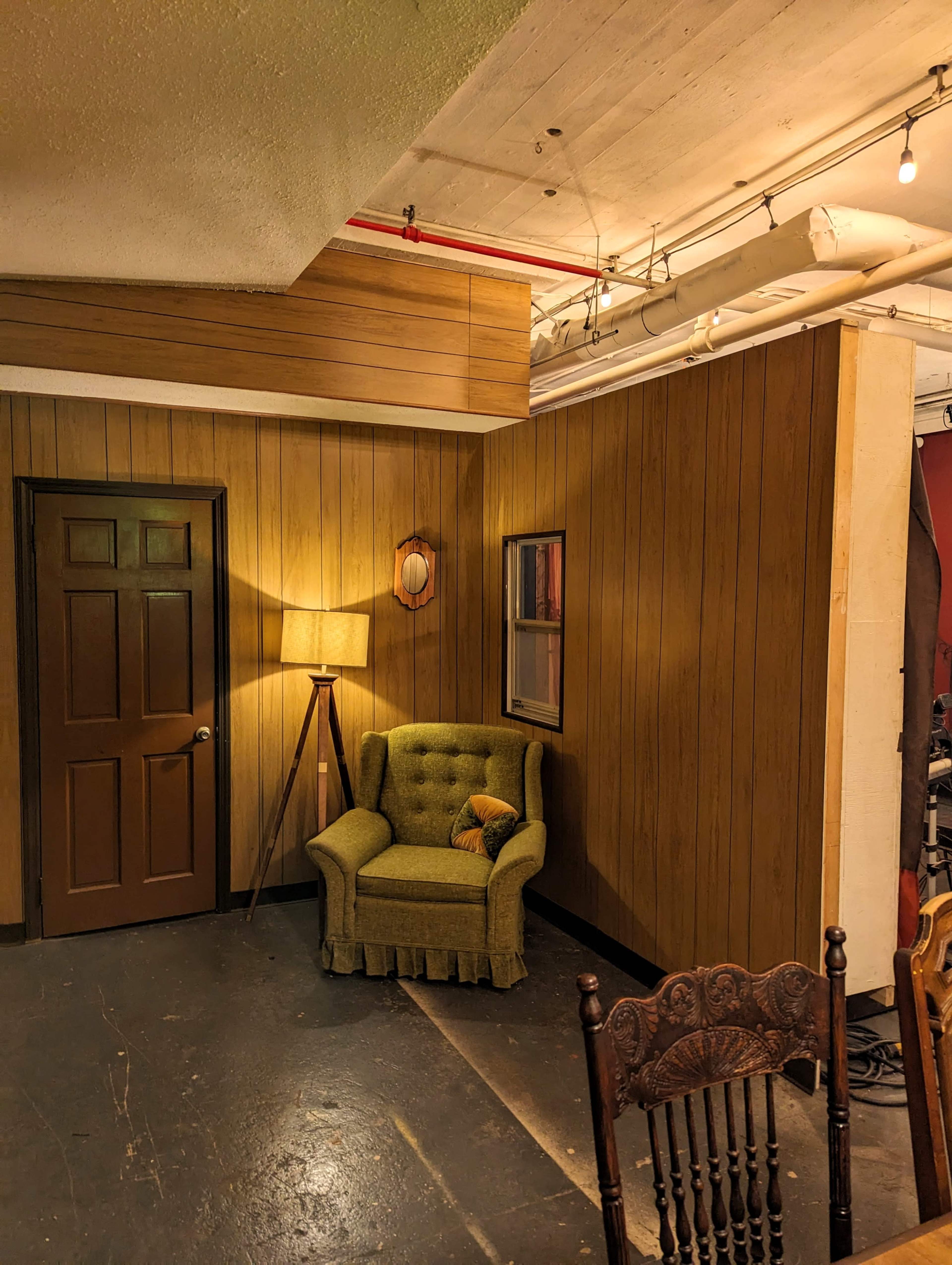 A small room with wooden paneling, featuring a green armchair, a floor lamp, a mirror on the wall, and a door.