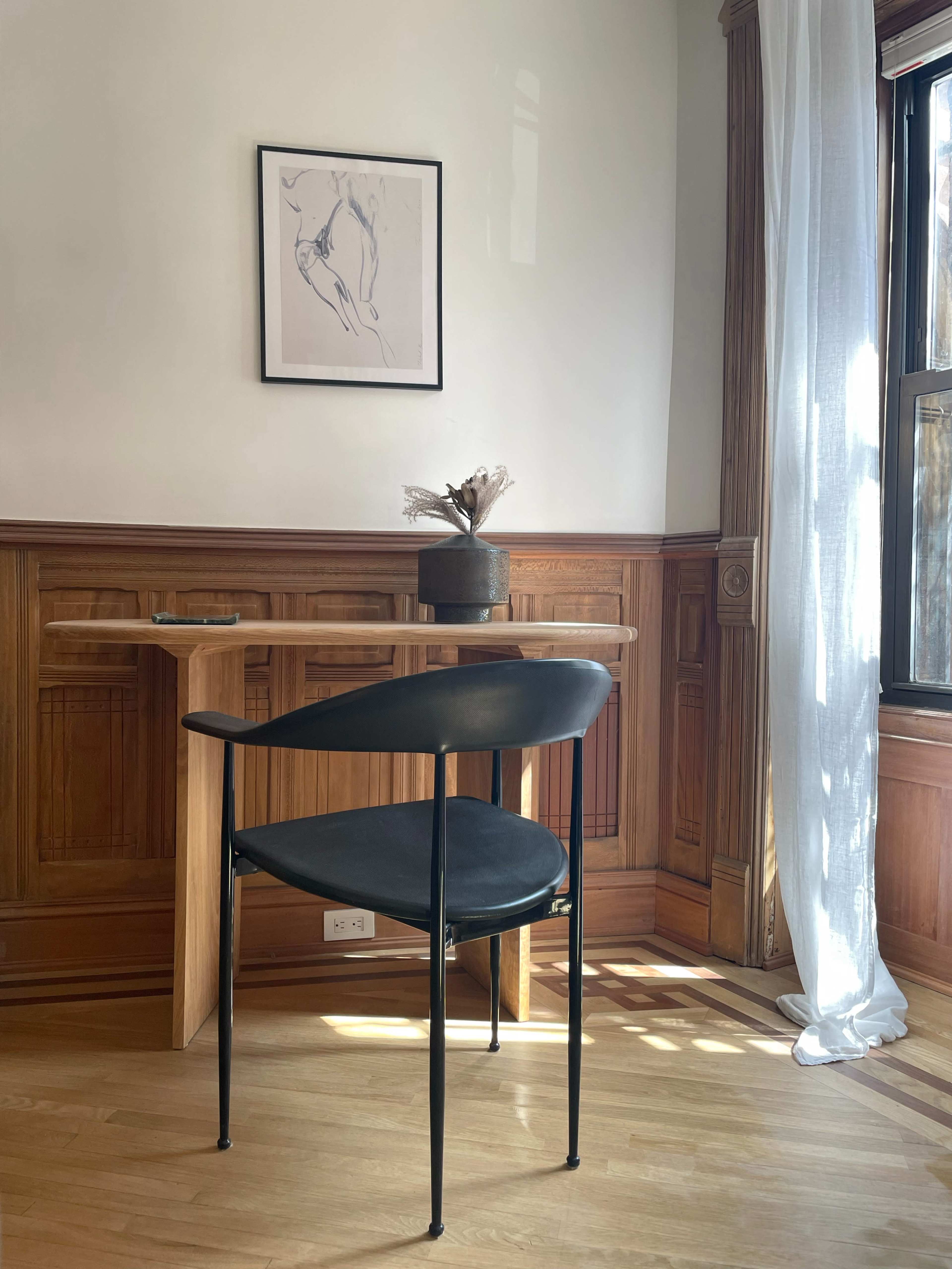 A chair is positioned beside a small wooden table with a decorative vase and a drawing on the wall in a sunlit room.