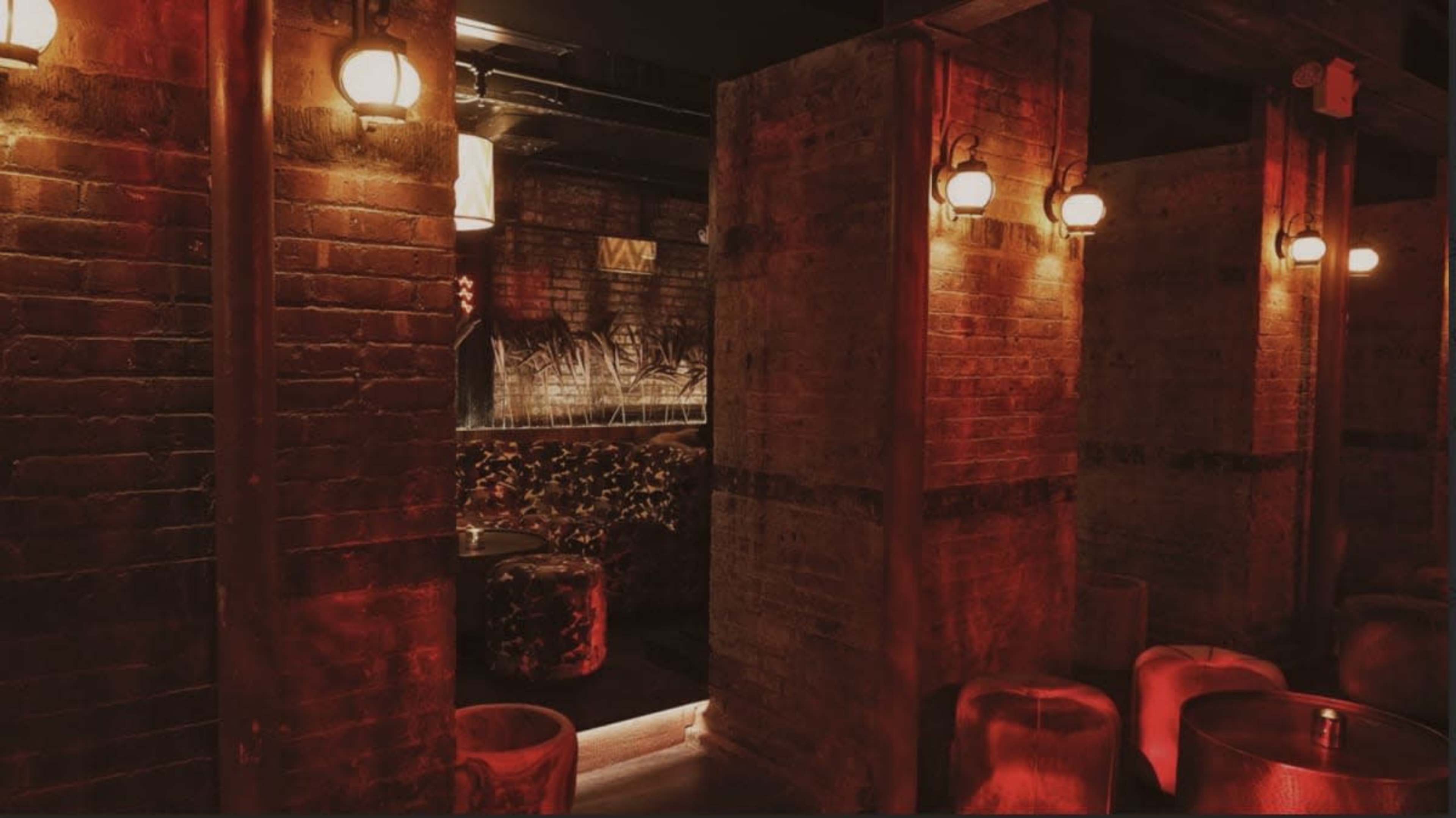 The image shows a dimly lit bar area with exposed brick walls and warm lighting, featuring several wooden tables and partitions.