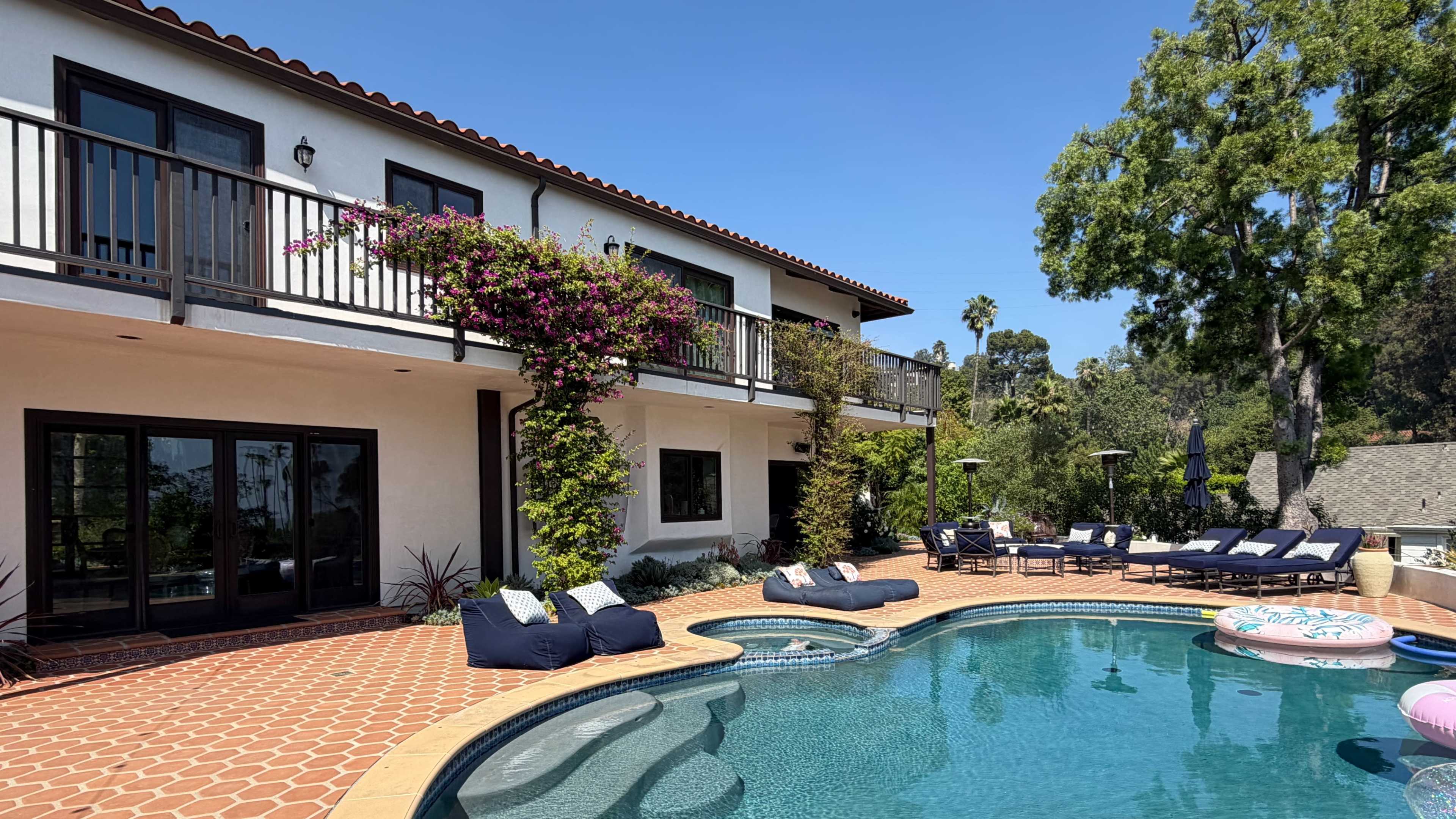 The image shows a modern home with a landscaped pool area featuring lounge chairs and vibrant greenery, set against a clear blue sky.