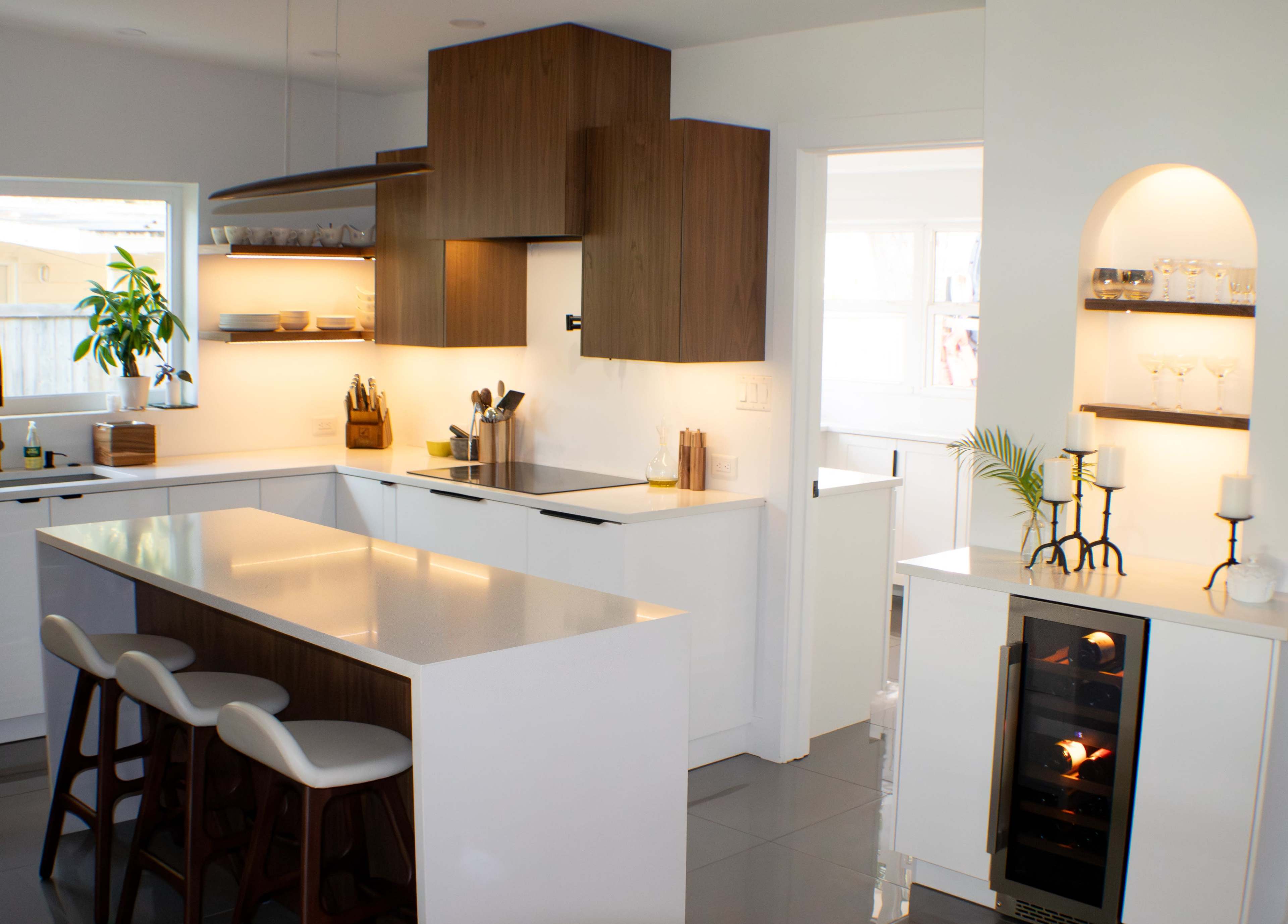 A modern kitchen features a central island with three stools, sleek cabinetry, and a wine cooler beside an arched nook with glassware.